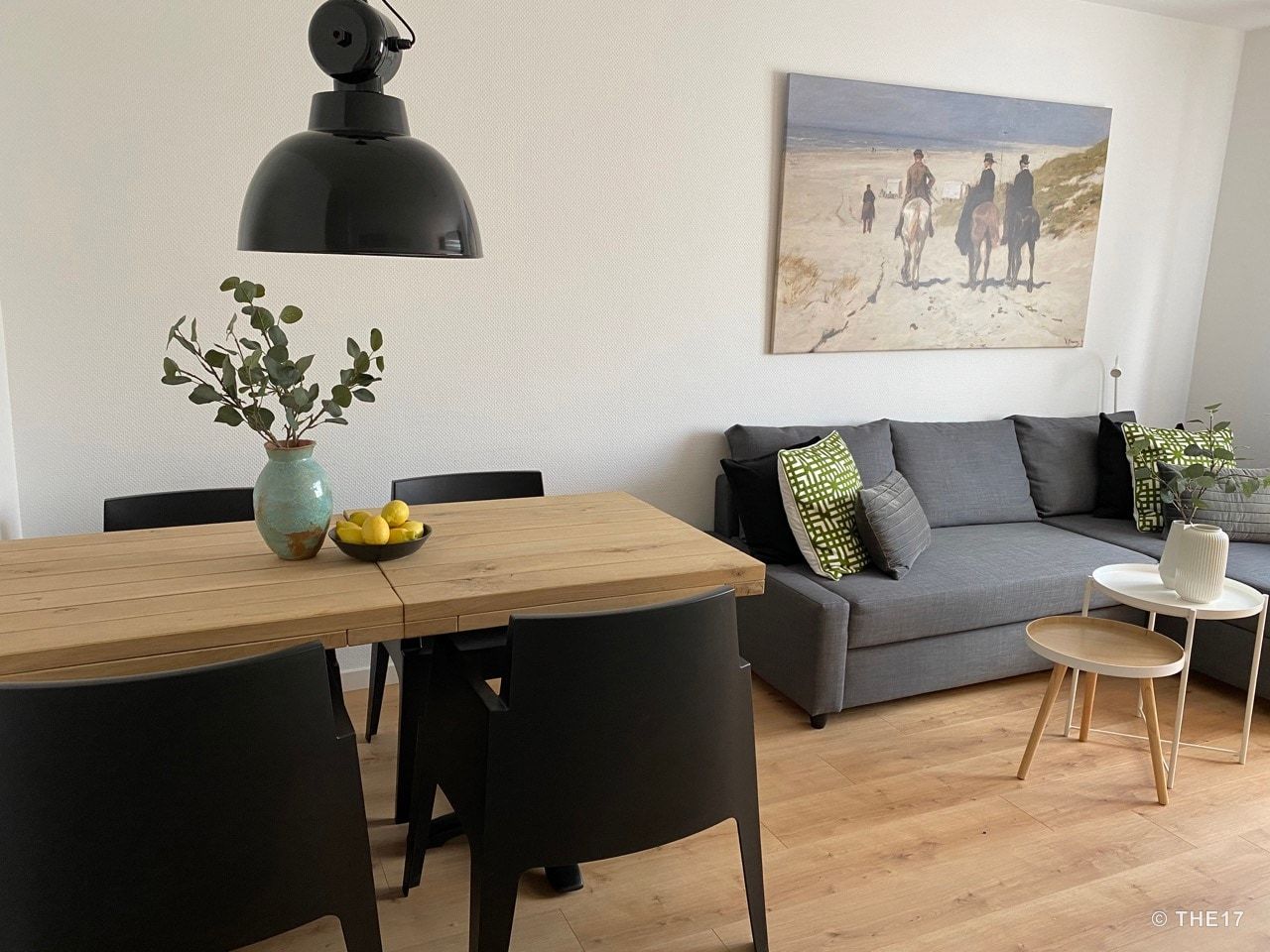 Moderner Essbereich mit Holztisch, schwarzen Stühlen, grauem Sofa und Wandbild einer Reiterszene am Strand.