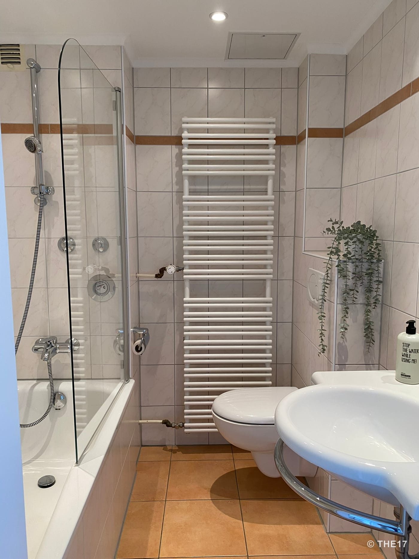 Kleines Badezimmer mit weißer Badewanne, Duschabtrennung aus Glas, wandhängendem WC, Heizkörper und Waschbecken mit Seifenspender.
