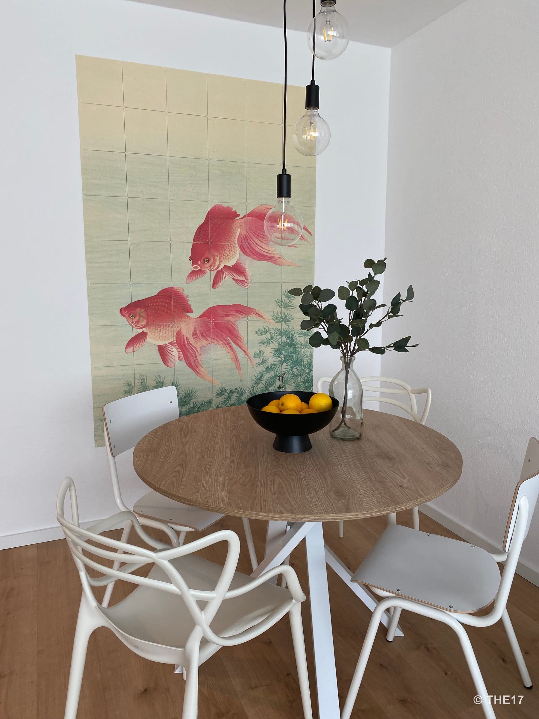 Moderner Essbereich mit runden Holztisch, weißen Stühlen, zwei hängenden Glühlampen, Vasen mit Grünpflanzen und Wandbild mit roten Fischen.