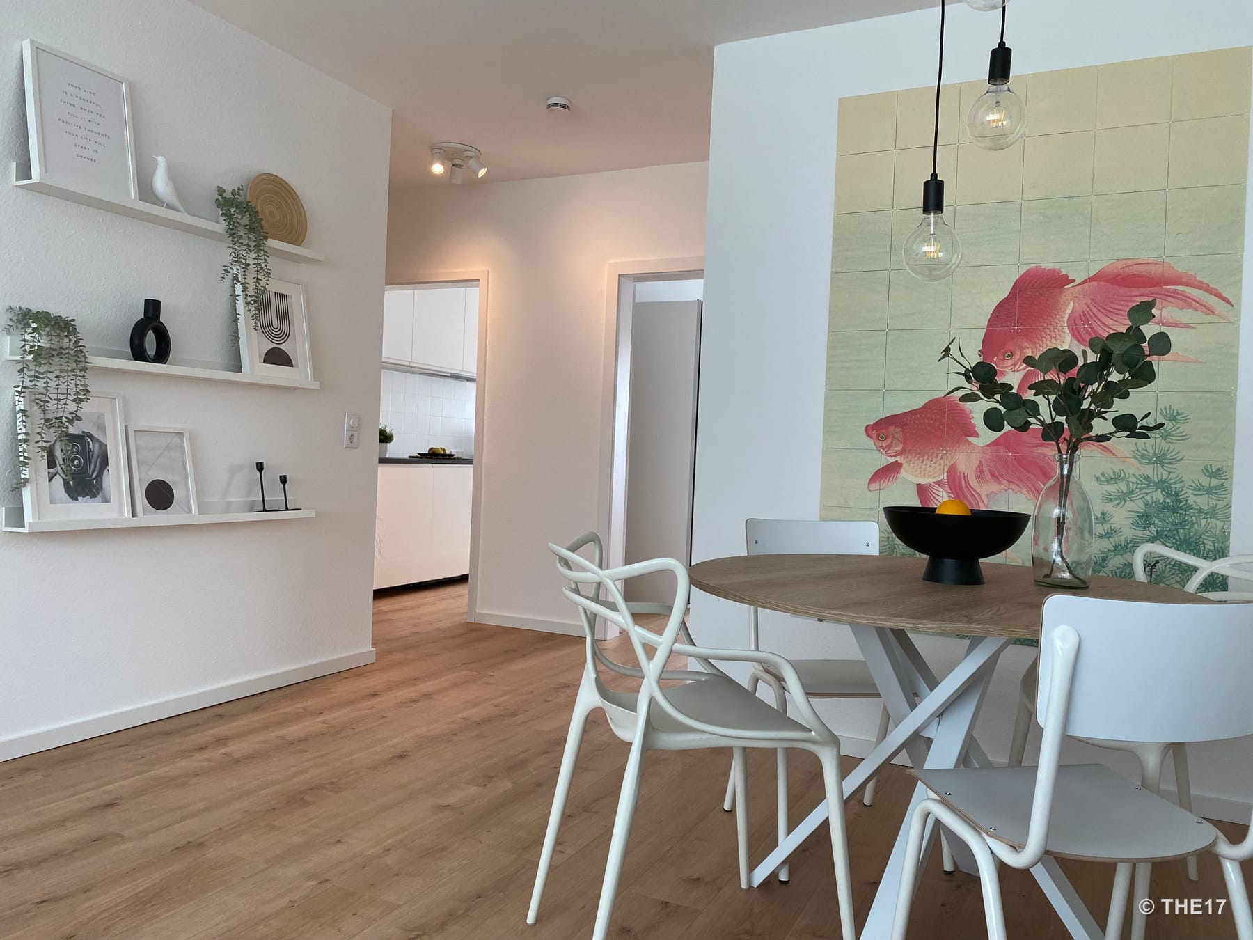 Essbereich mit rundem Holztisch, weißen Stühlen, großen Wandbildern und offener Küche mit weißen Schränken im Hintergrund.