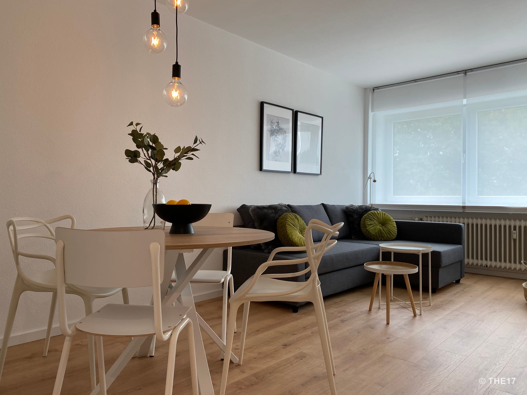 Essbereich mit rundem Holztisch, weißen Stühlen, grauem Sofa mit grünen Kissen und großen Fenstern mit halbtransparenten Rollos.