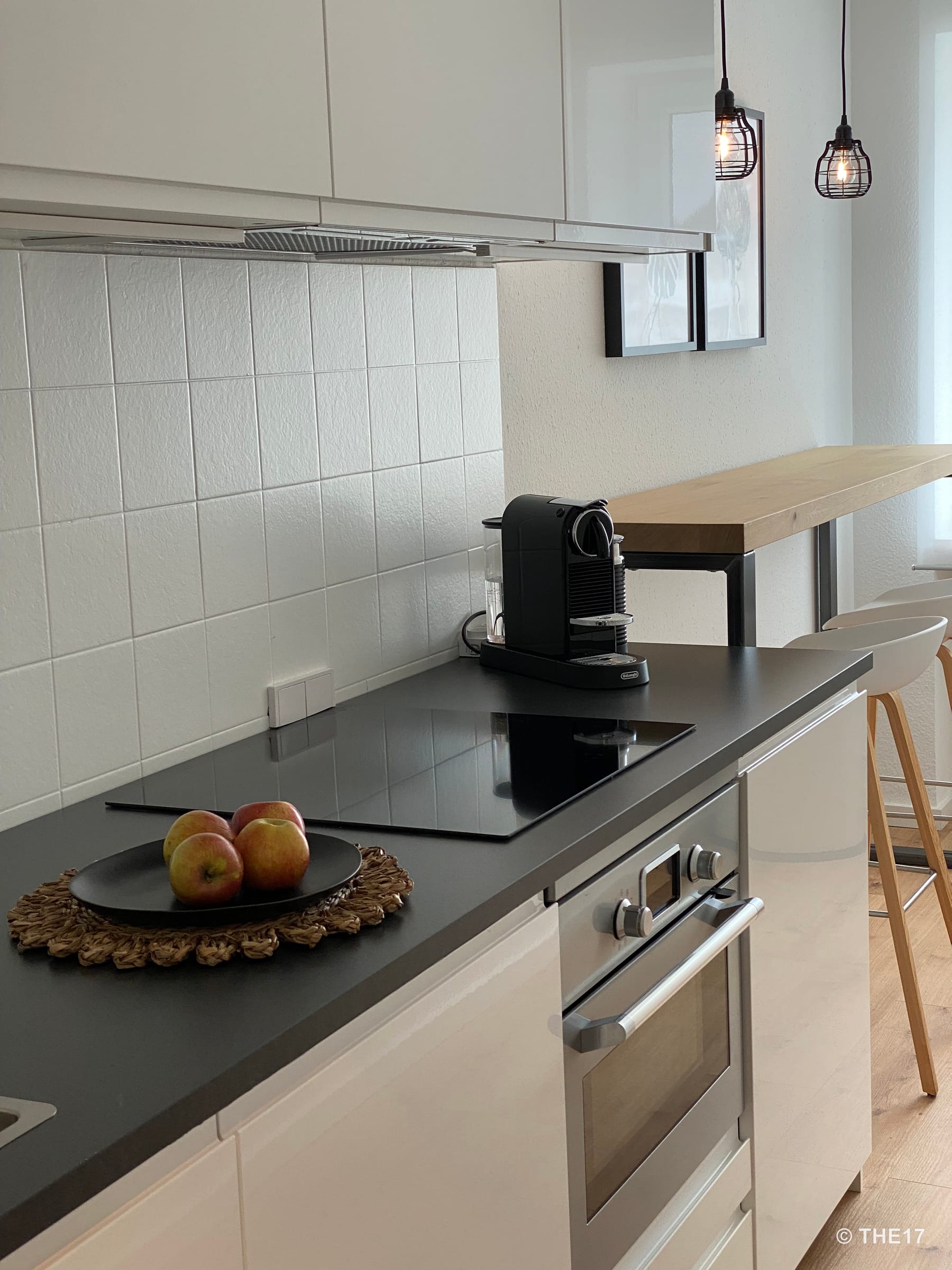 Moderne Küche mit schwarzer Arbeitsplatte, Cerankochfeld, Einbauofen, Kaffeemaschine und Holz-Bar mit Hockern.