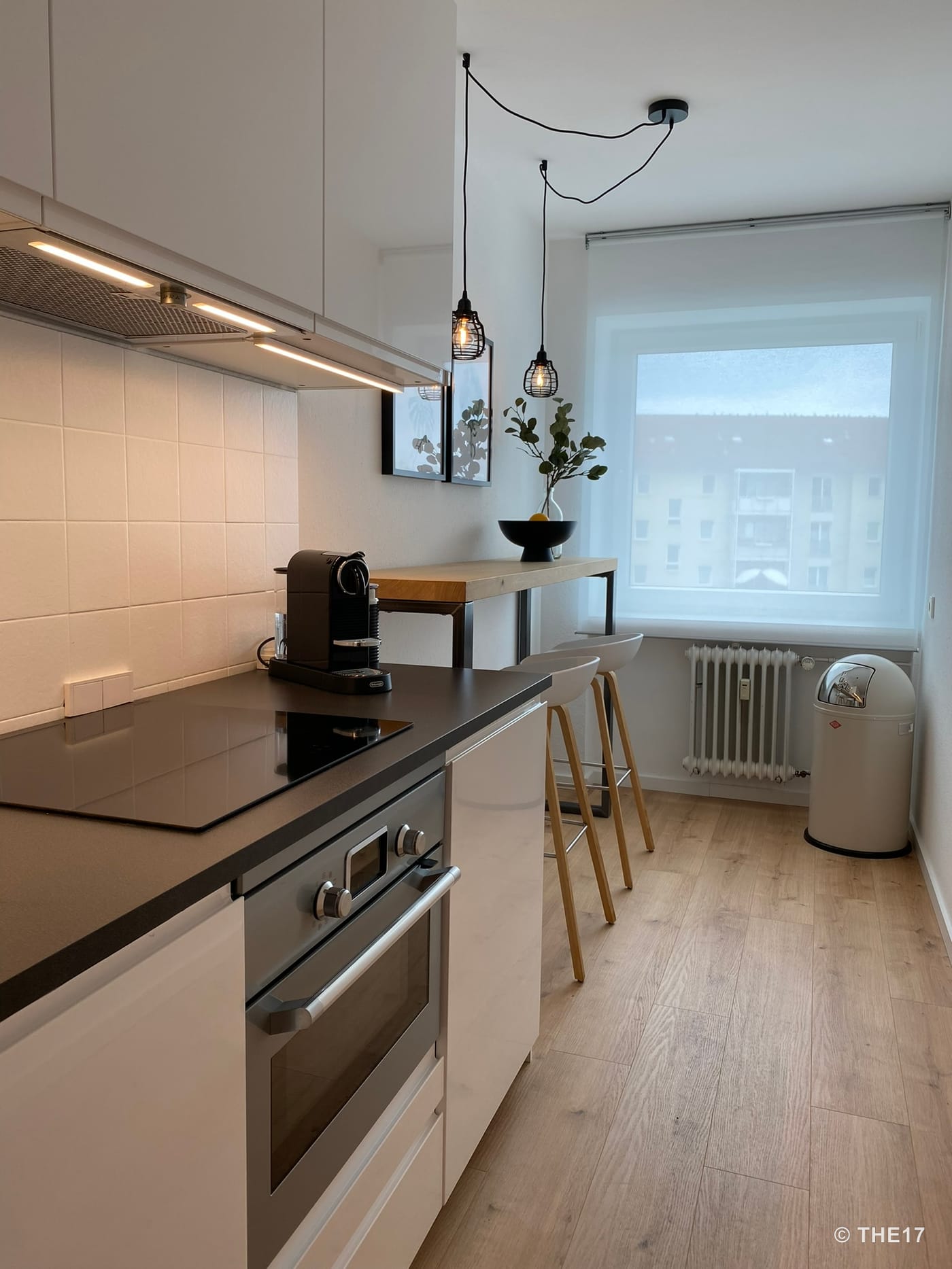 Helle, schmale Küche mit Holzfußboden, schwarzer Arbeitsplatte, Herd, Kaffeemaschine, Tresen mit zwei Hockern und großem Fenster.