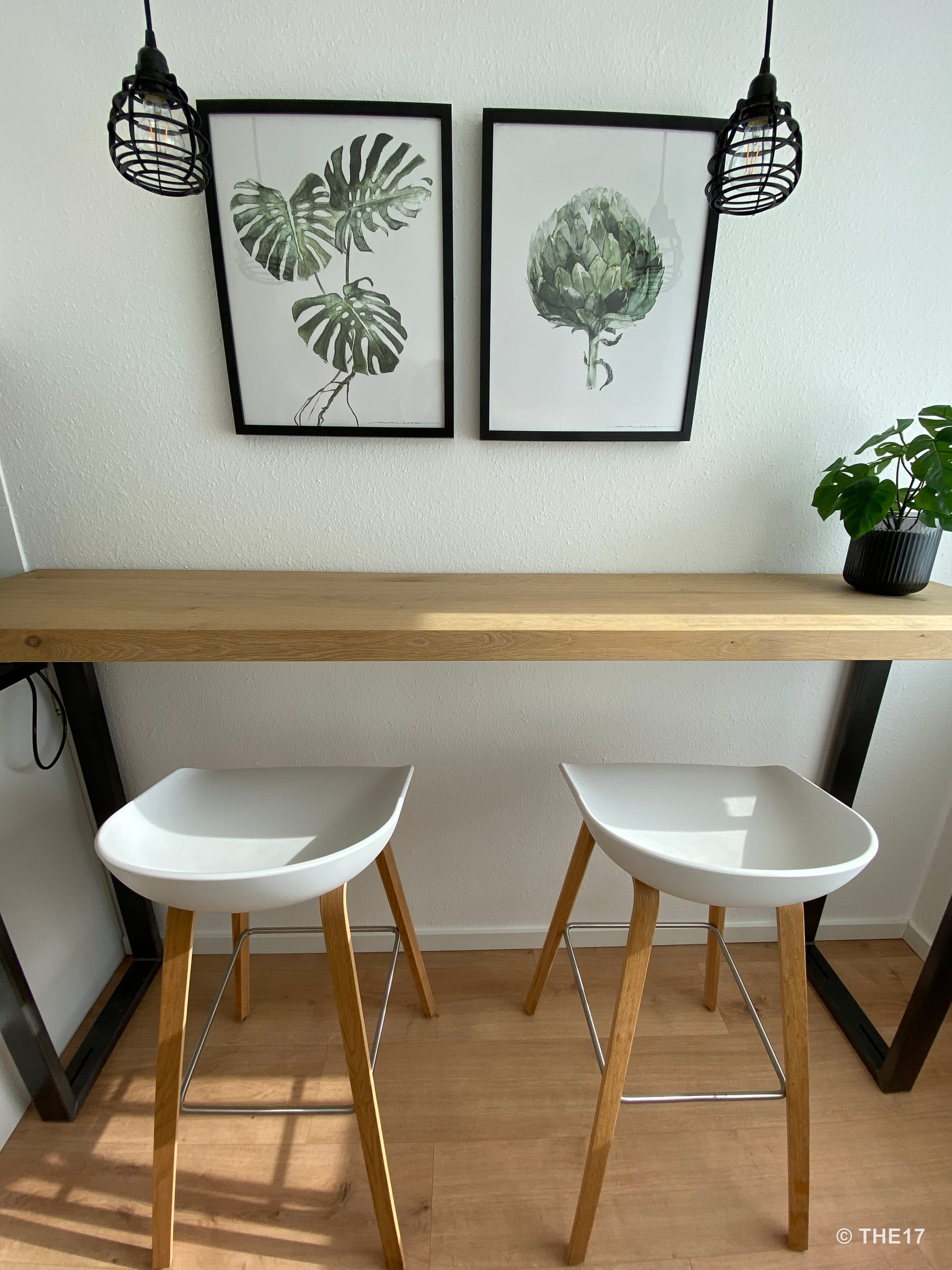 Zwei weiße Barhocker mit Holzbeinen vor einer Holztheke, darüber zwei Bilder mit grünen Pflanzenmotiven und zwei schwarze Hängeleuchten.