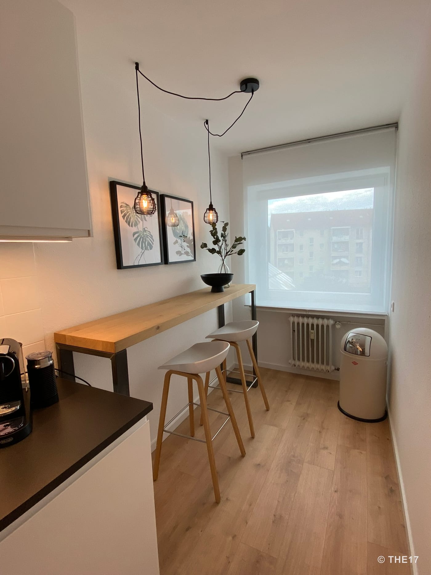 Schmale Küche mit Holztheke, zwei Barhockern, zwei hängenden Lampen, zwei Bilderrahmen und großem Fenster mit Sicht auf Gebäude.