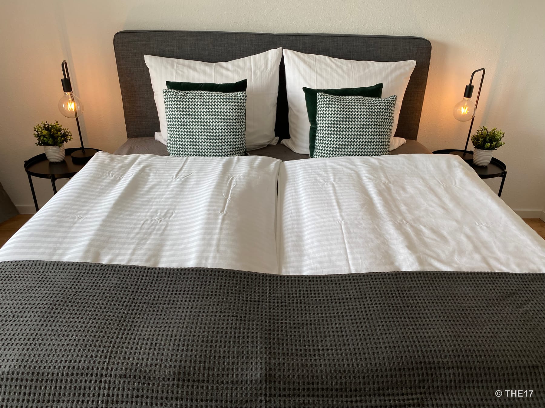 Doppelbett mit grauem Kopfteil, weißen Kissen und Decken, grünen Zierkissen, zwei schwarzen Nachttischen mit Lampen und Pflanzen.