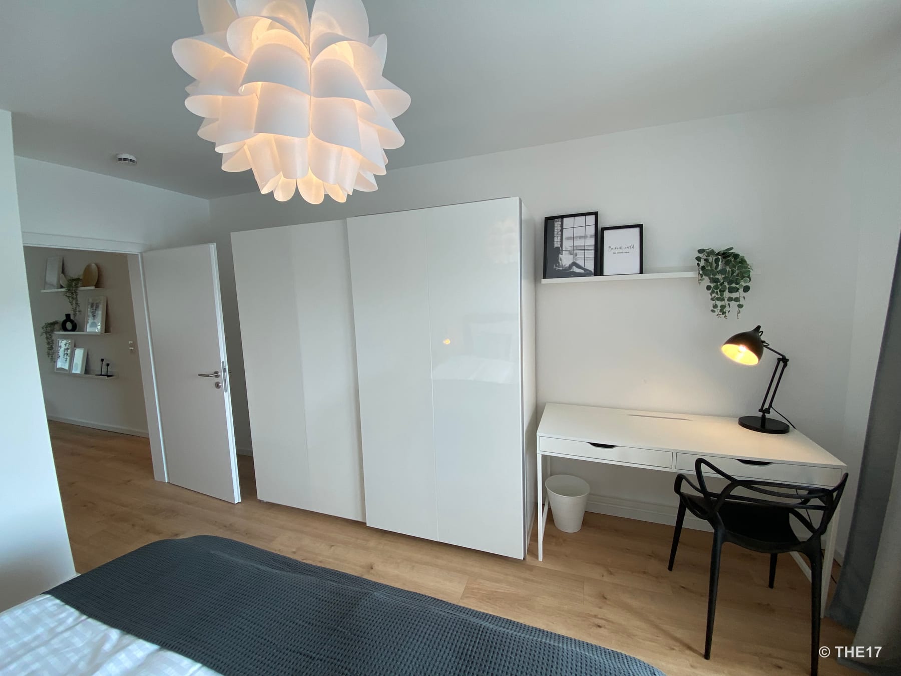 Schlafzimmer mit weißem Kleiderschrank, Schreibtisch mit Lampe, schwarzem Stuhl und großem Bett mit dunkelgrauer Decke.