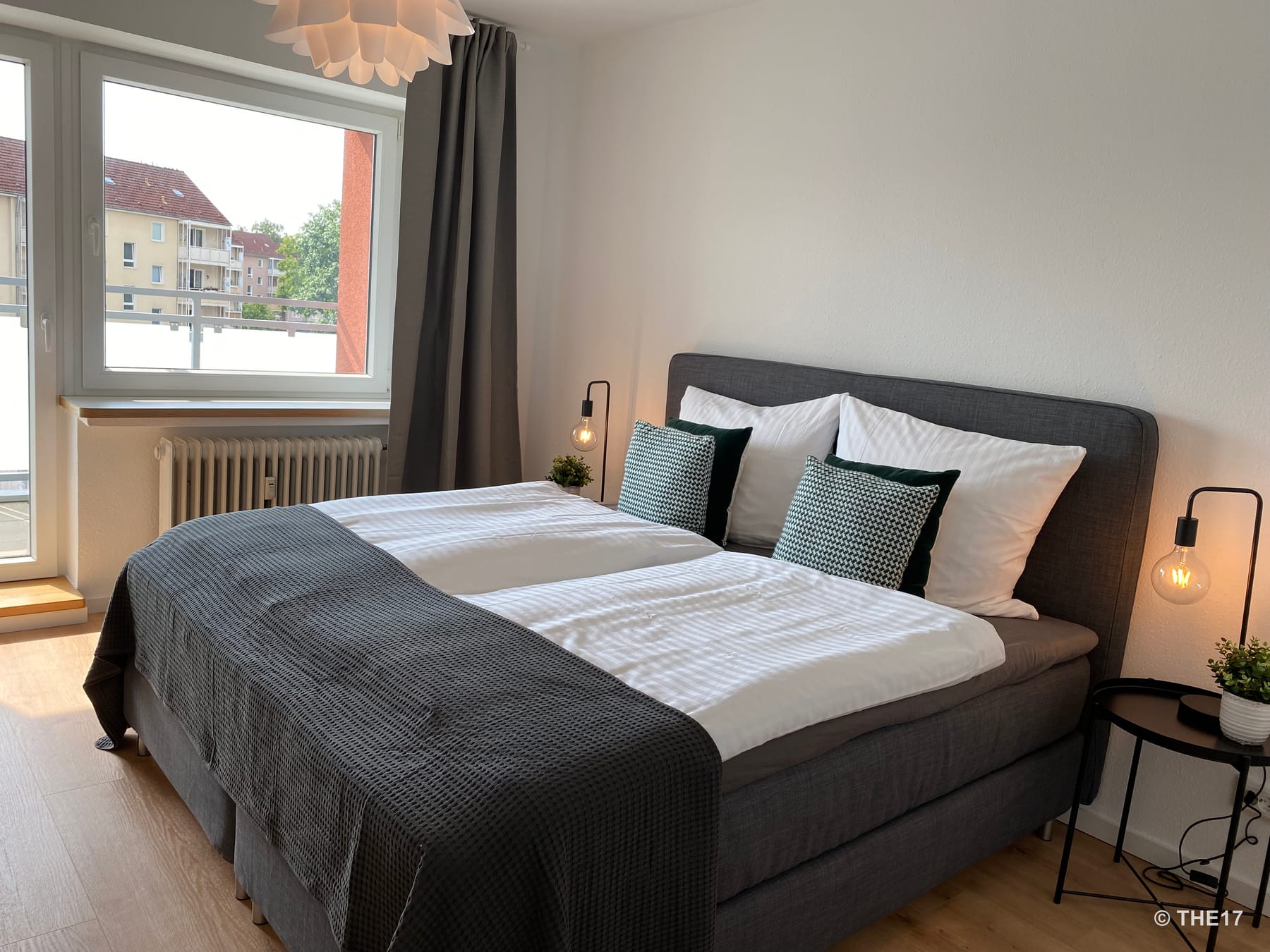 Modernes Schlafzimmer mit großem Doppelbett, weißen und gemusterten Kissen, grauer Bettdecke und bodentiefem Fenster mit Vorhang.