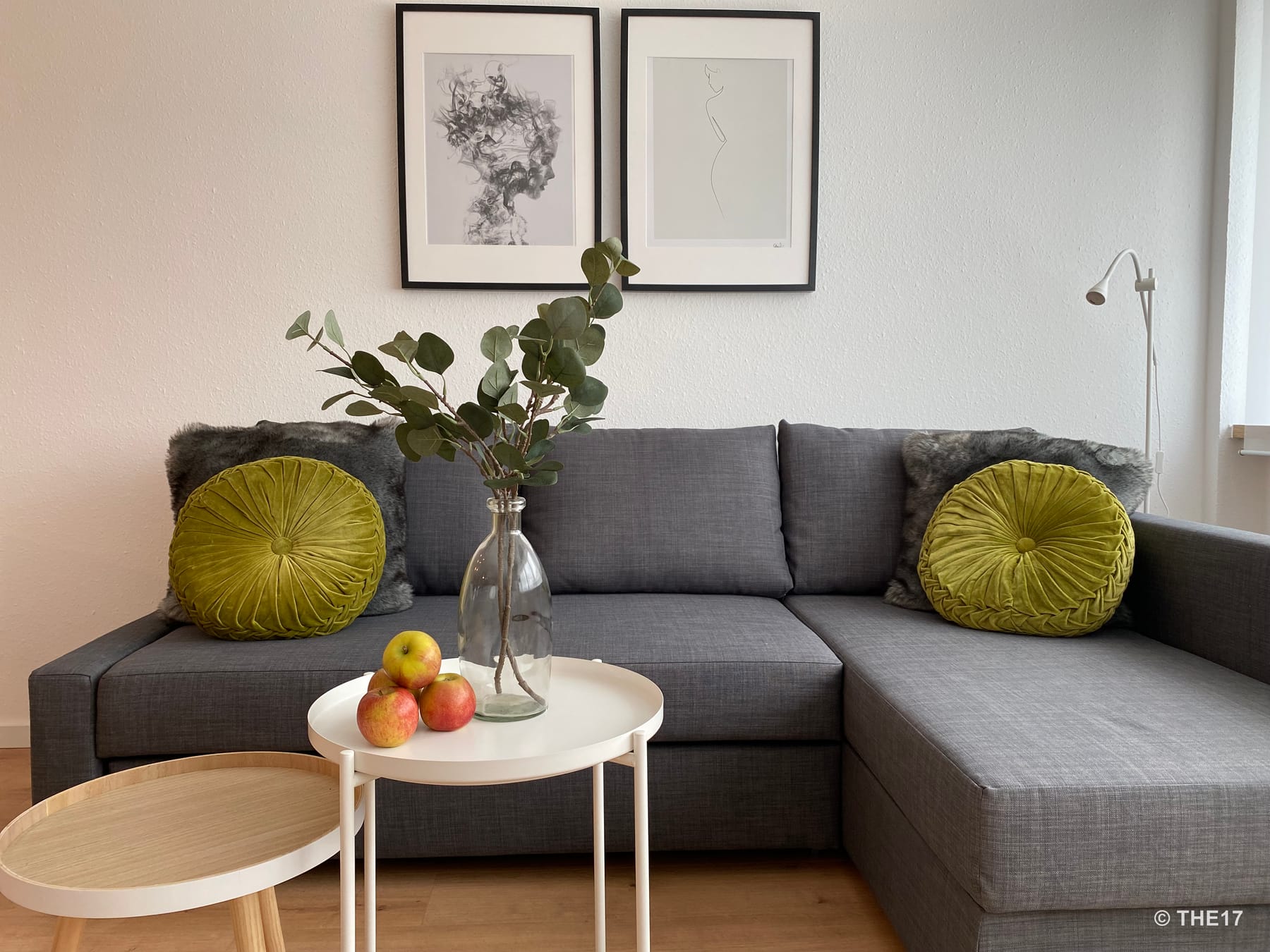 Graues Ecksofa mit grünen runden Kissen und grauen Fellen, zwei Tischen mit Vase und Äpfeln, minimalistische Bilder an der Wand.