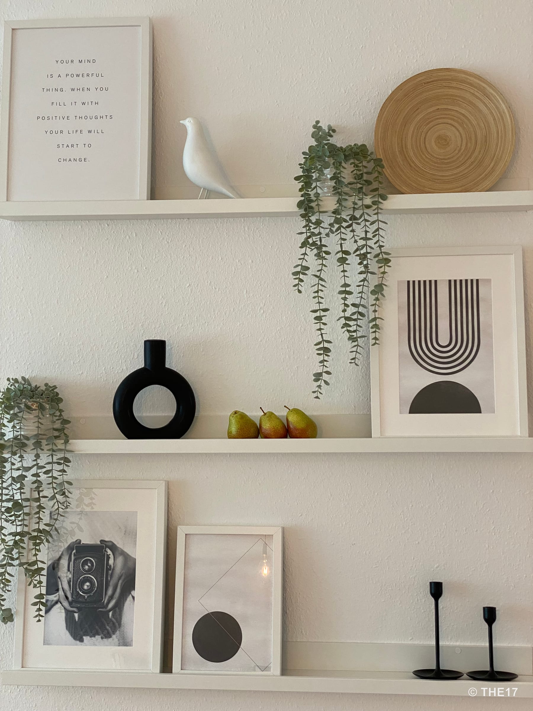 Weiße Wandregale mit Bildern, einer schwarzen Vase, künstlichen Pflanzen, drei Birnen und Kerzenhaltern an weißer Wand.