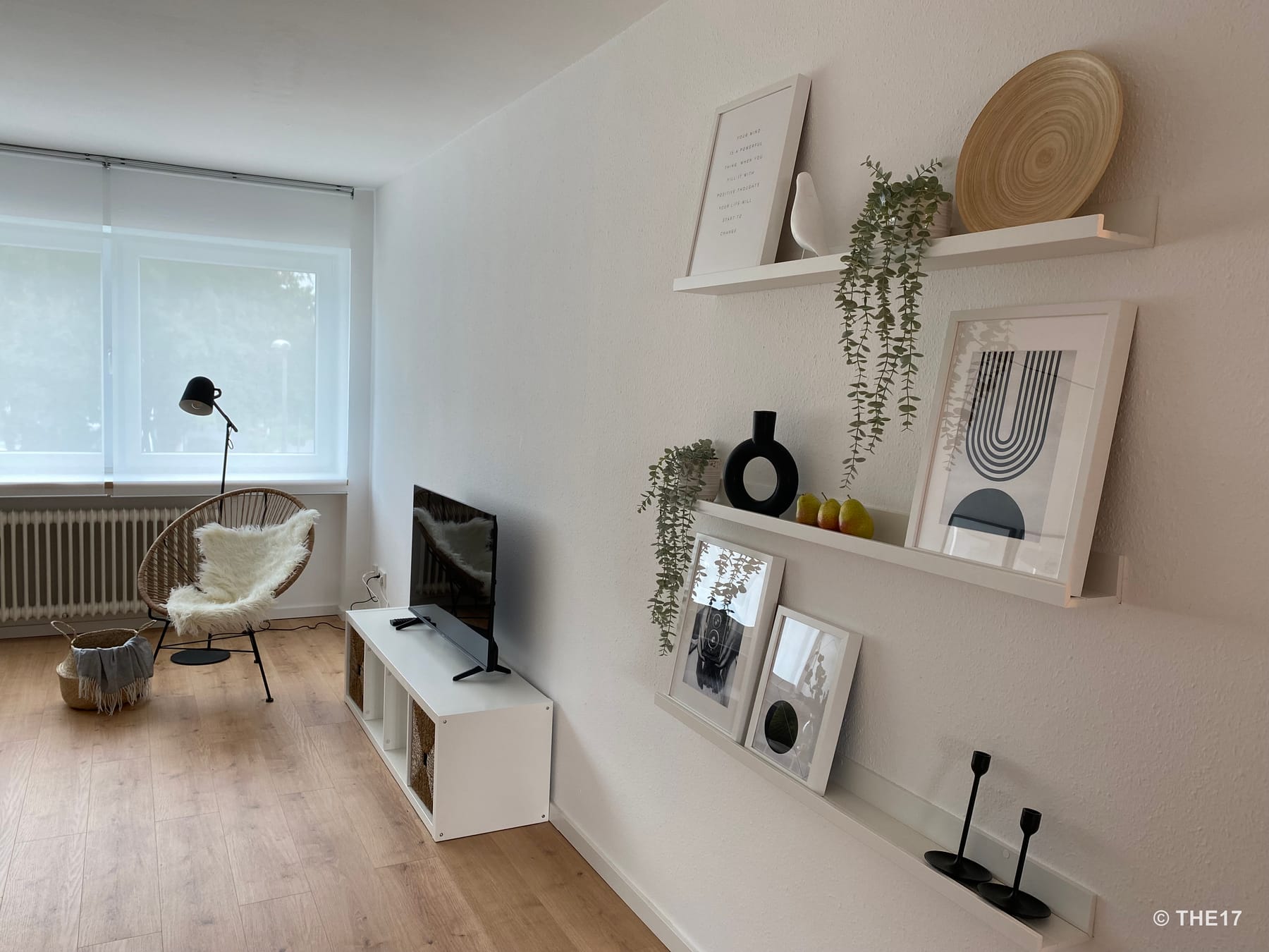 Minimalistisches Wohnzimmer mit Holzfußboden, TV auf weißem Regal, Rattanstuhl mit Fellvorleger und dekorativen Regalen an der Wand.