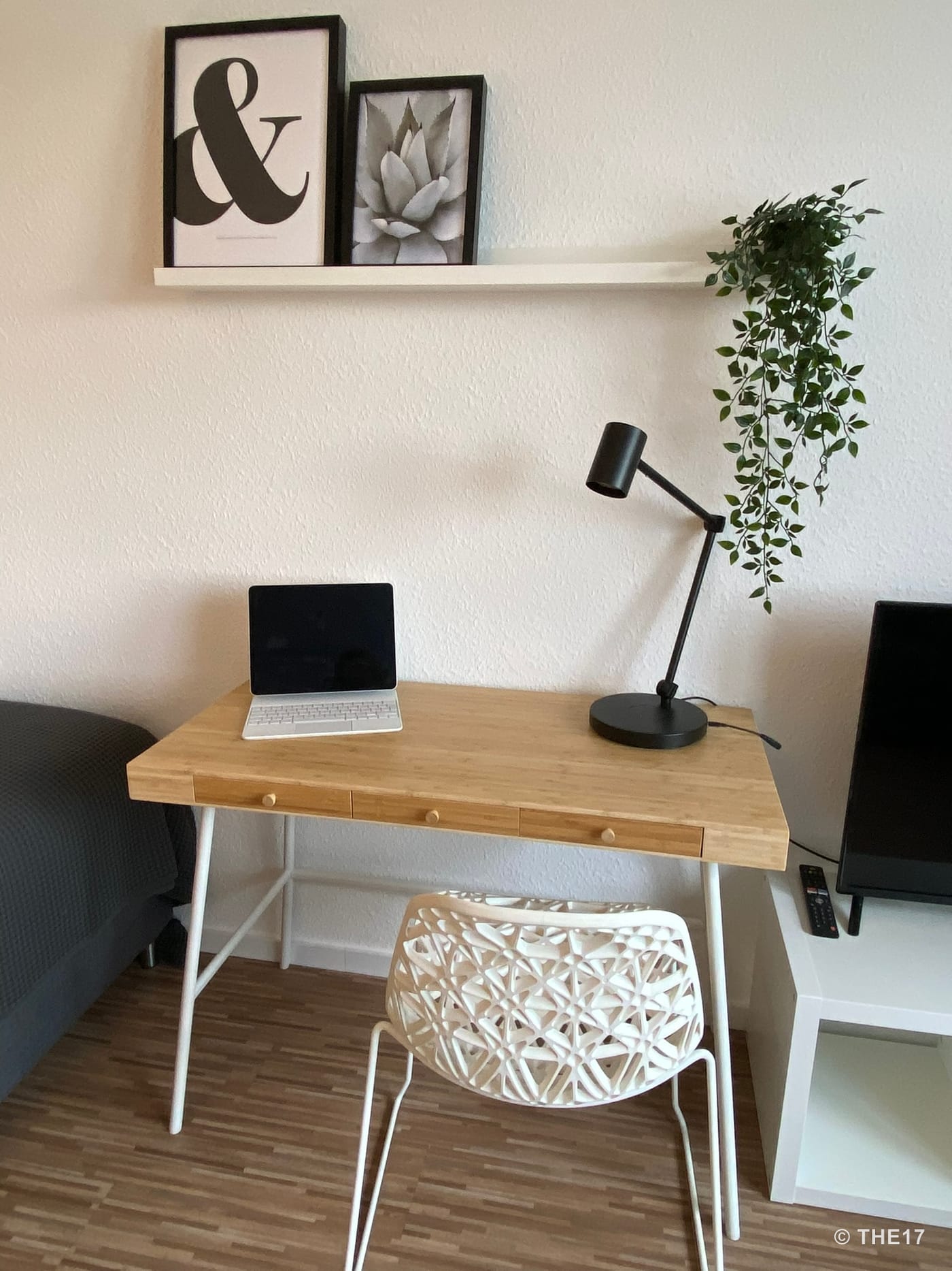 Modern workspace with wooden desk, white chair, black desk lamp, tablet, wall shelf with frames, and hanging plant.