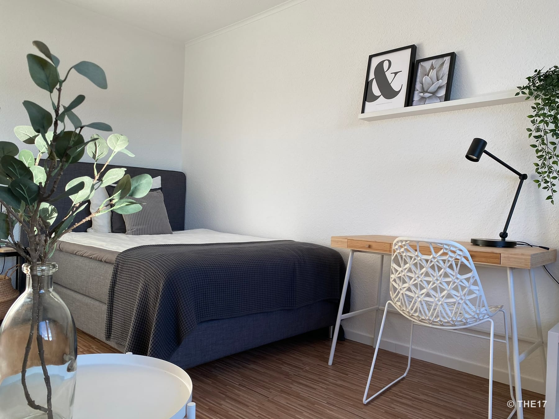 Bedroom with grey bed, dark blanket, small wooden desk with white chair and black desk lamp, plants in the room.