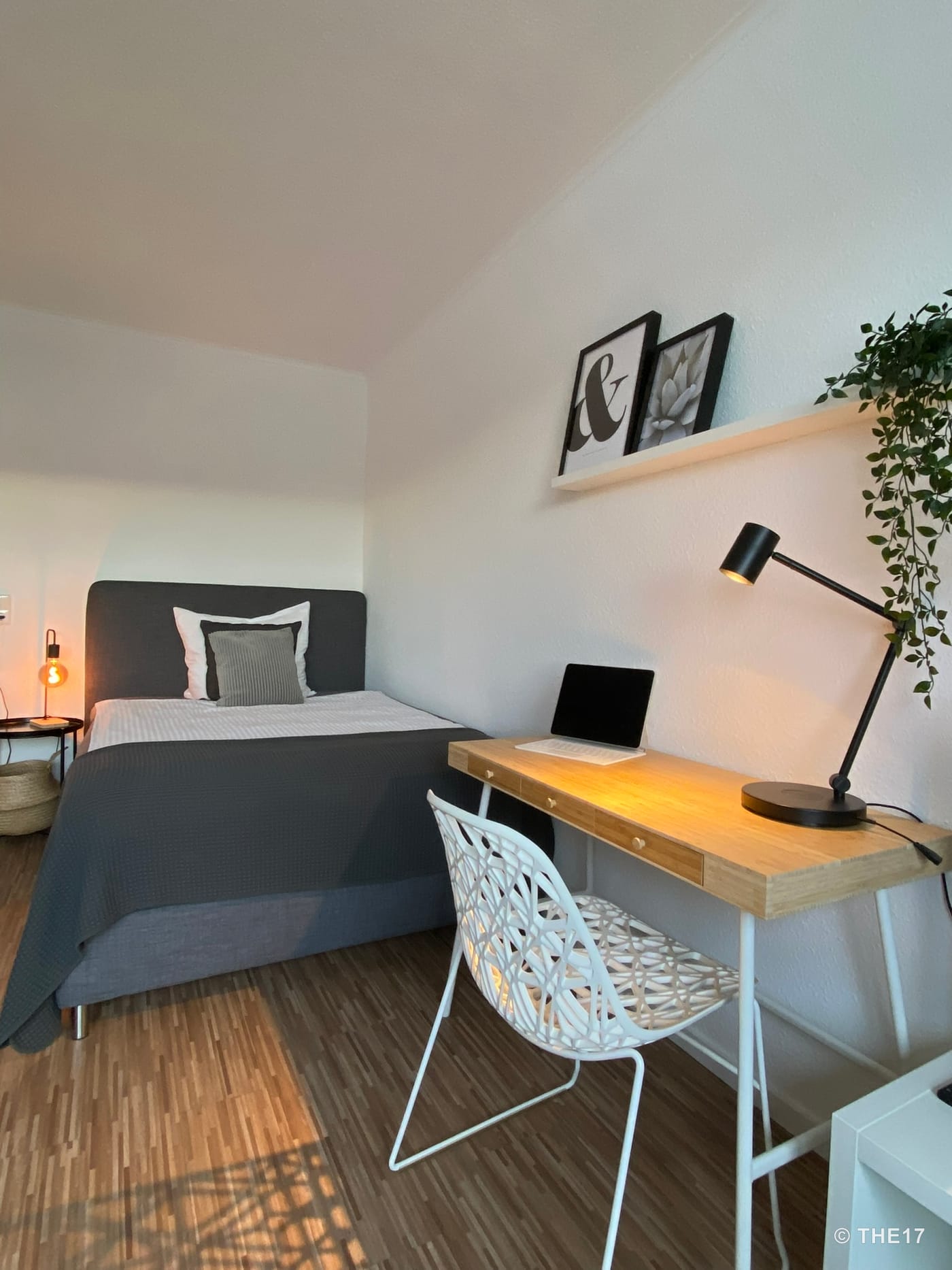 Small bedroom with gray bed, wooden desk, black lamp, white chair, and plant on the wall.