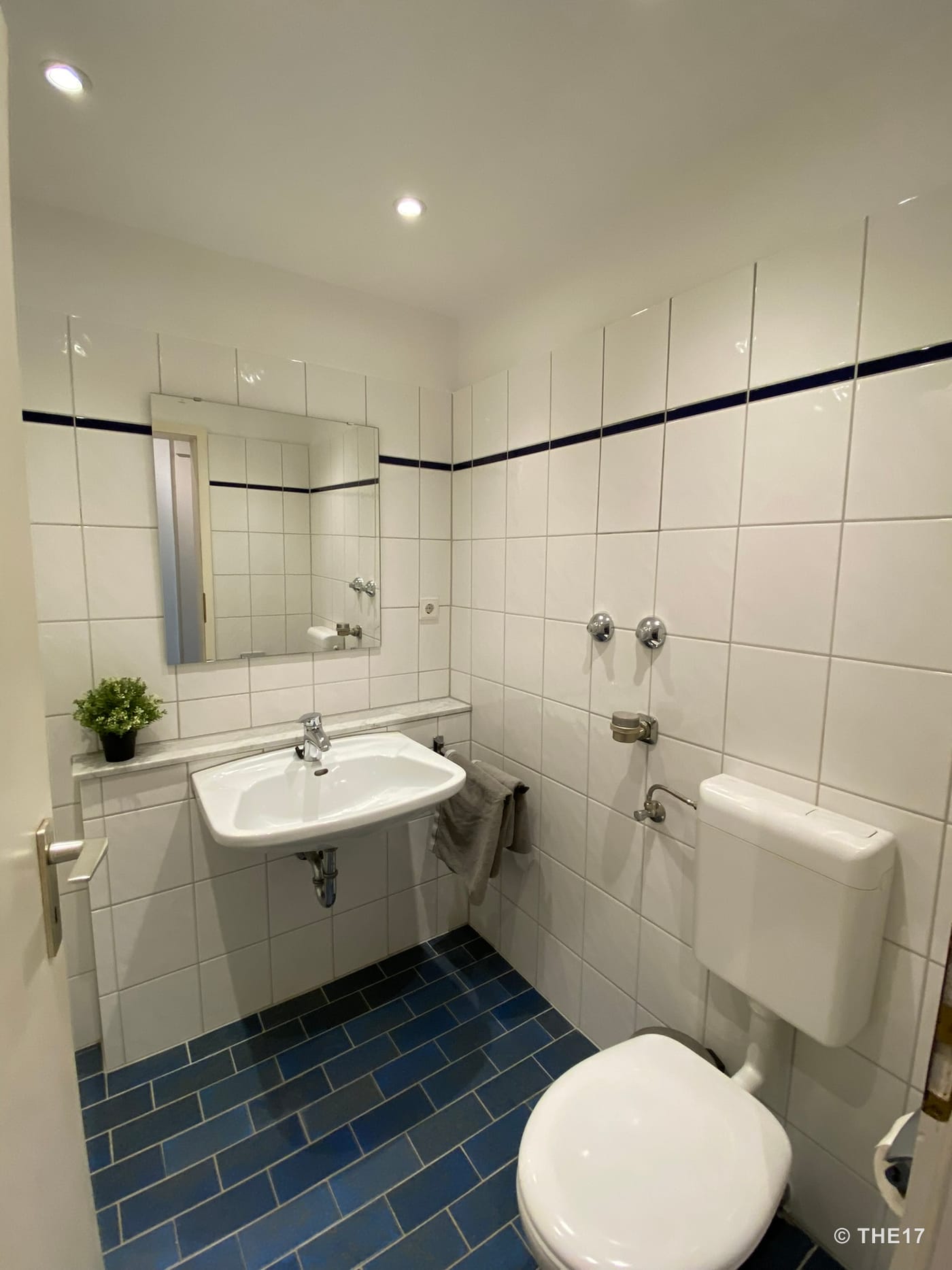 Small bathroom with white tiled walls, blue tiled floor, sink, mirror and toilet.