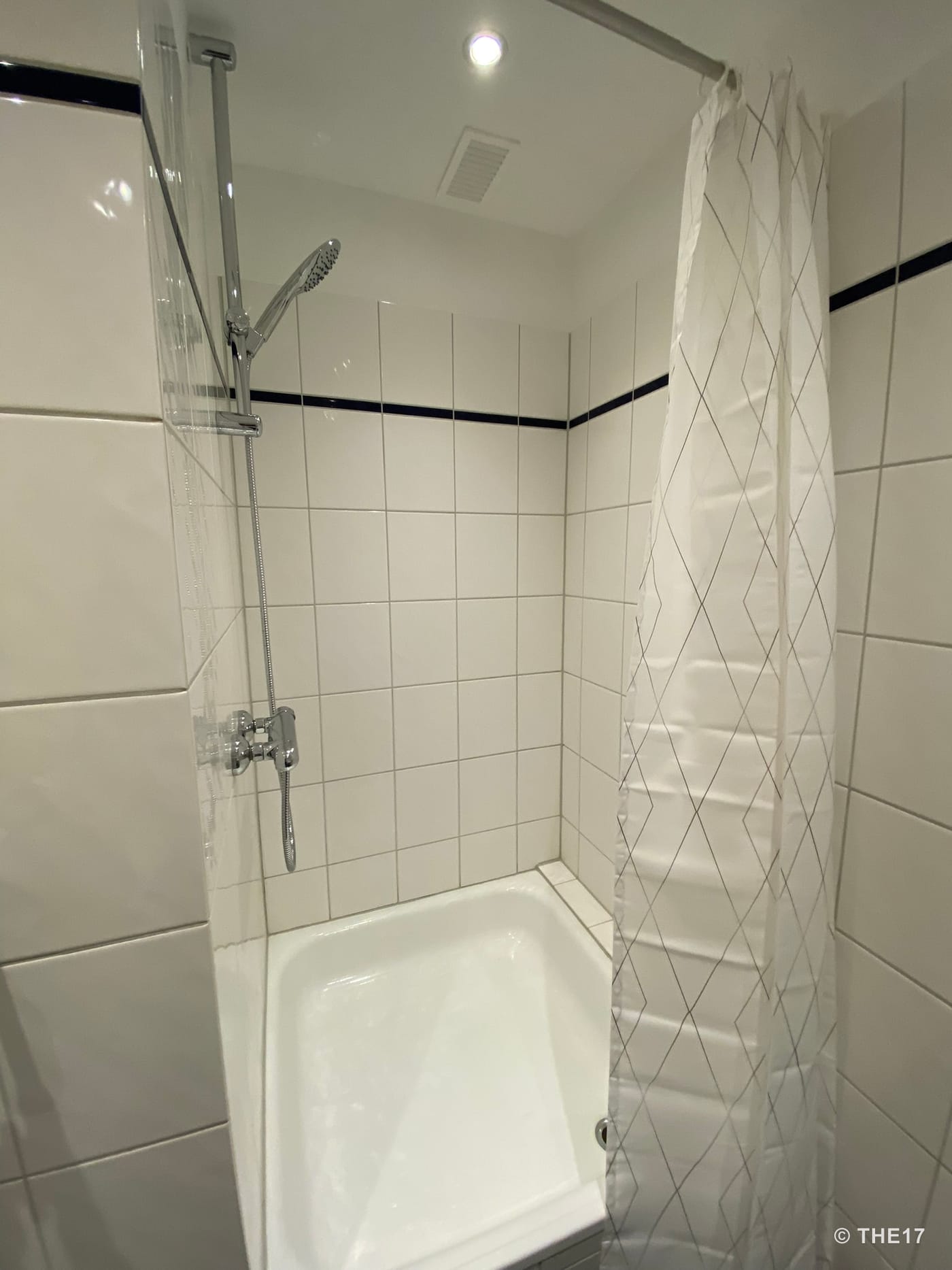 Small shower stall with white tiles, adjustable shower head, and white patterned shower curtain.
