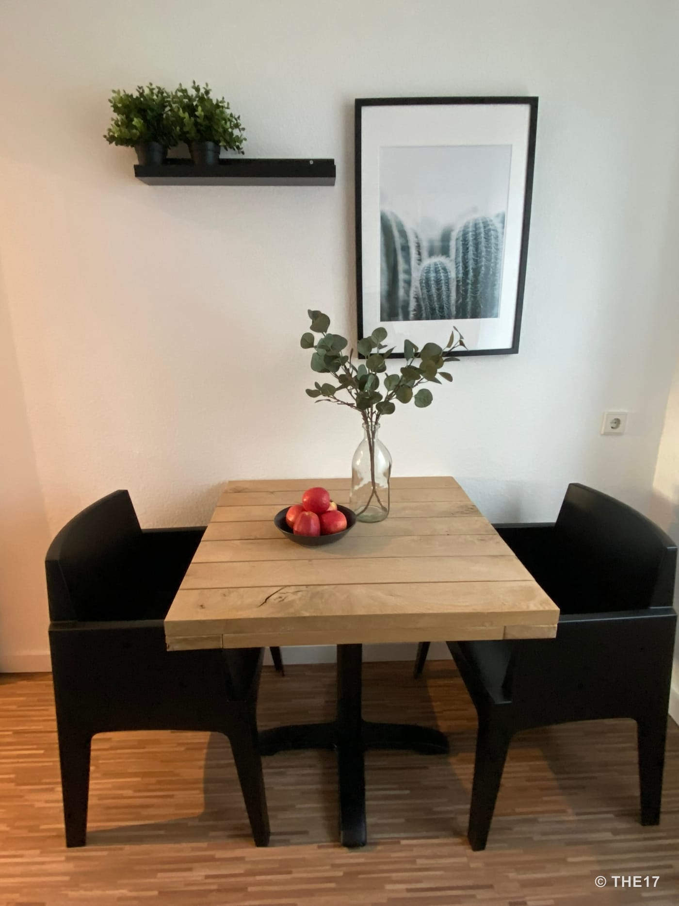 Small wooden table with two black chairs, a vase with branches and a bowl of red apples, wall art and shelf with plants.