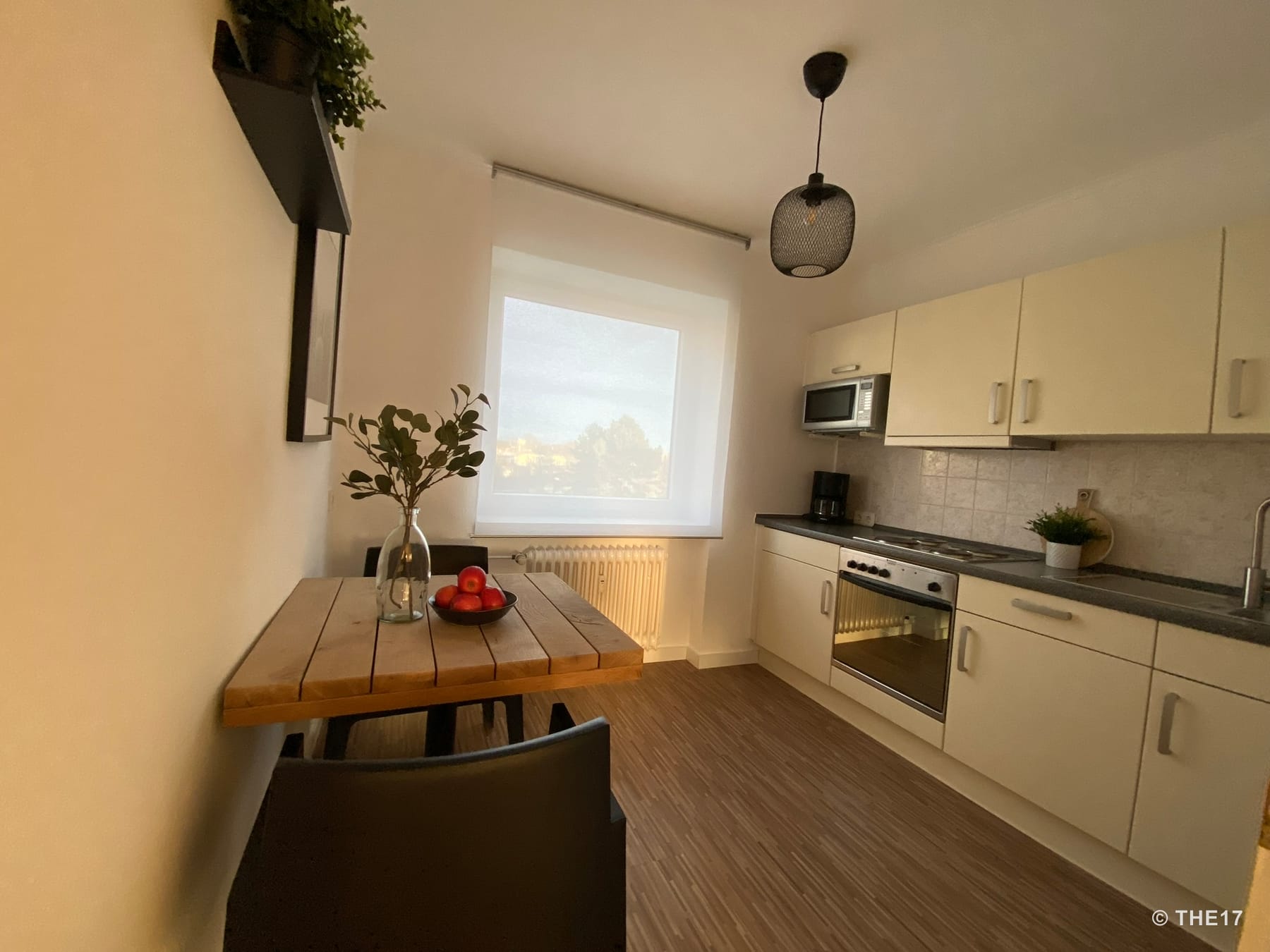 Small kitchen with wooden table, black chairs, white cabinets, oven, microwave, and window with white curtain.