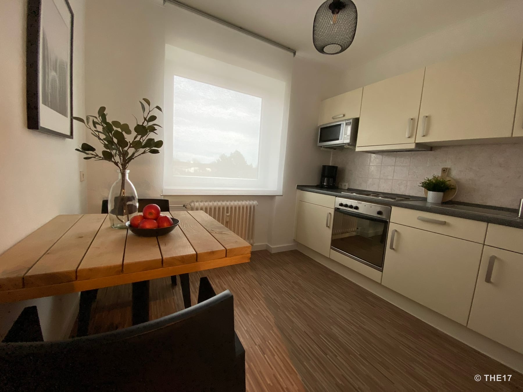 Small kitchen with wooden table, black chairs, white curtain, stove, microwave, and decoration with apples and plants.