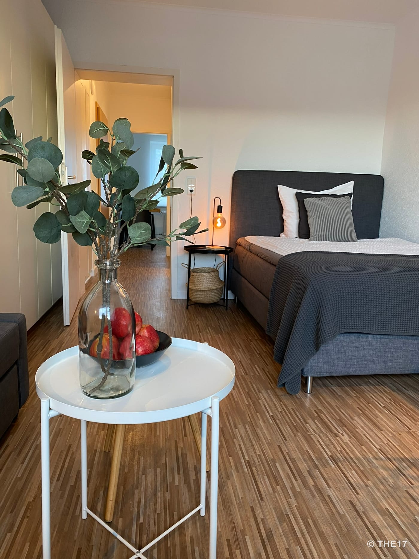 Bedroom with gray bed, white nightstand, hanging light bulb, white round table with glass vase and red apples, wooden floor.