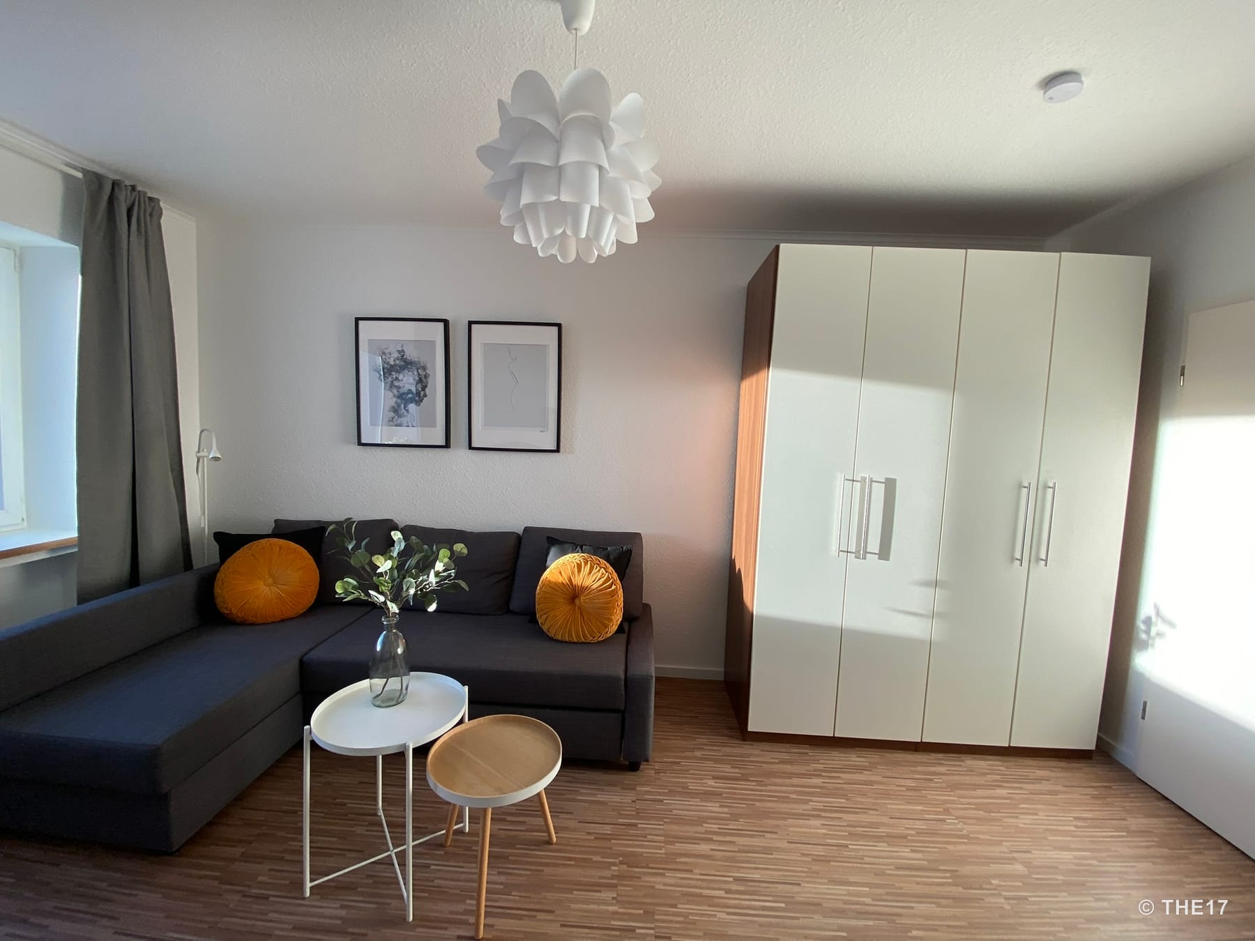 Cozy room with dark gray corner sofa, yellow round cushions, two small tables, white wardrobe, and two framed pictures on white wall.