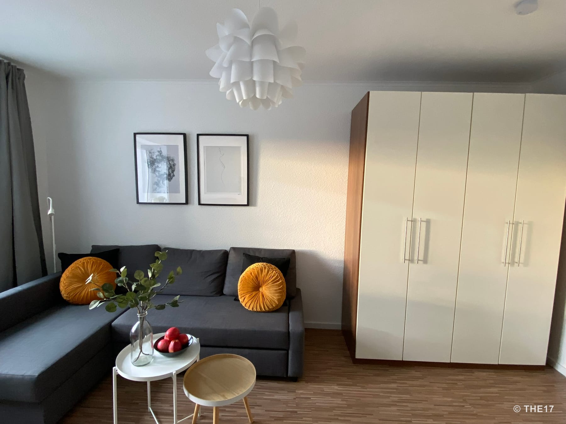 Living room with gray sectional sofa, yellow round cushions, two tables with decorations, large white wardrobe, and two framed pictures.
