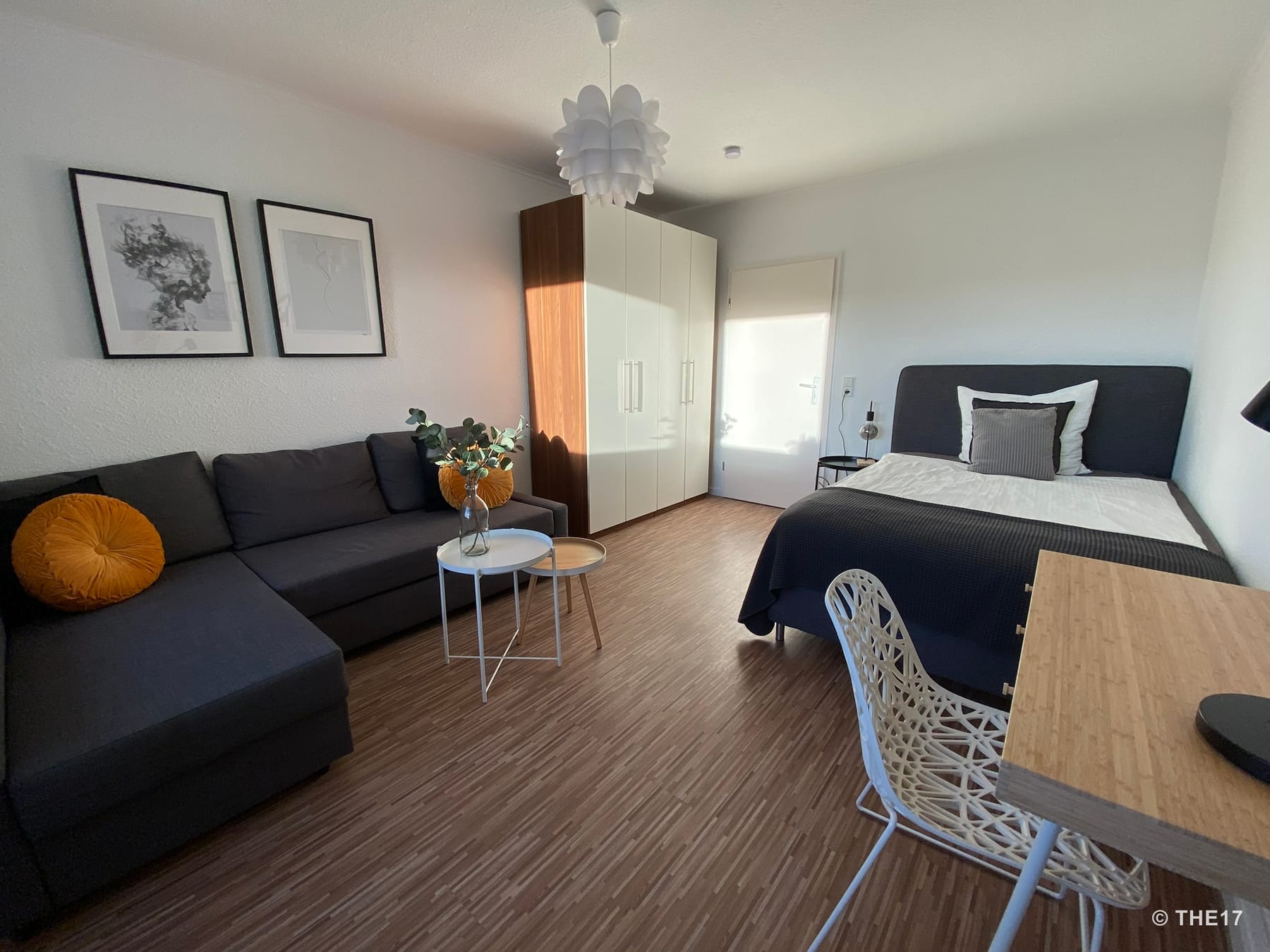 Bright room with gray sofa, two yellow cushions, white wardrobe, double bed with dark bedding, and desk with chair.