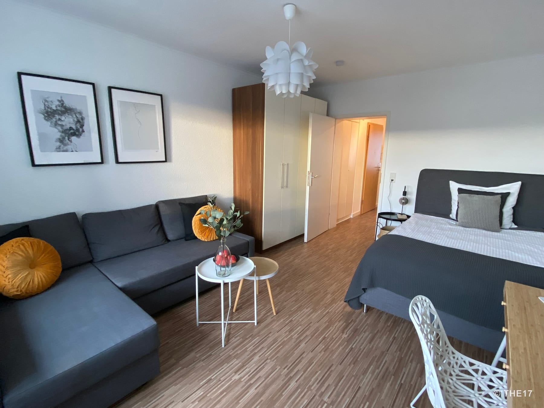 Room with gray sectional sofa, two round tables, double bed, wooden wardrobe, and white designer chair on wood laminate floor.