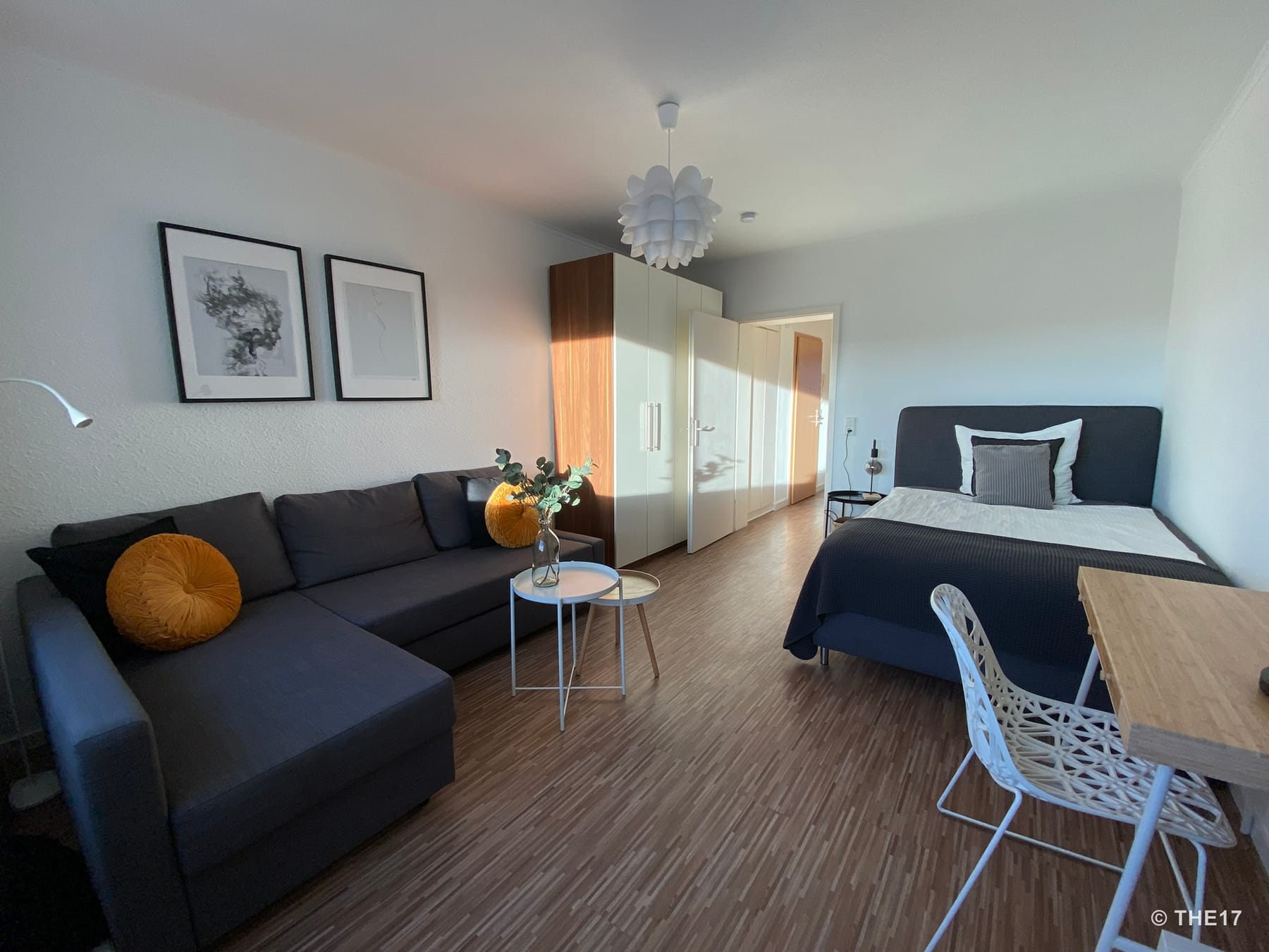 Bedroom with double bed, gray sectional sofa with orange cushions, two small tables, wardrobe, and desk with chair.