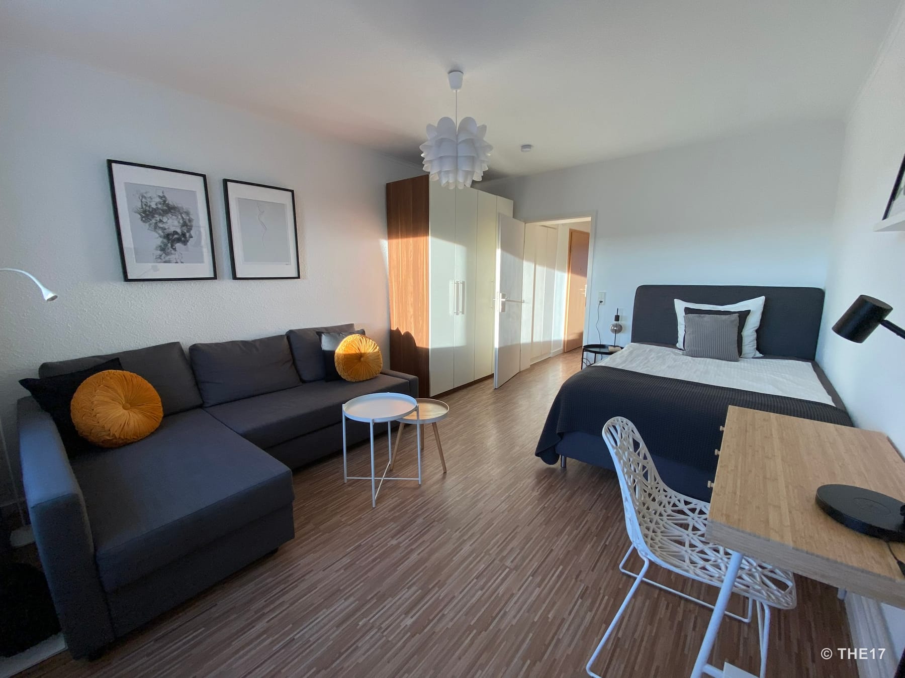 Living room with dark gray sectional sofa and orange cushions, wardrobe, double bed, and desk with white chair.