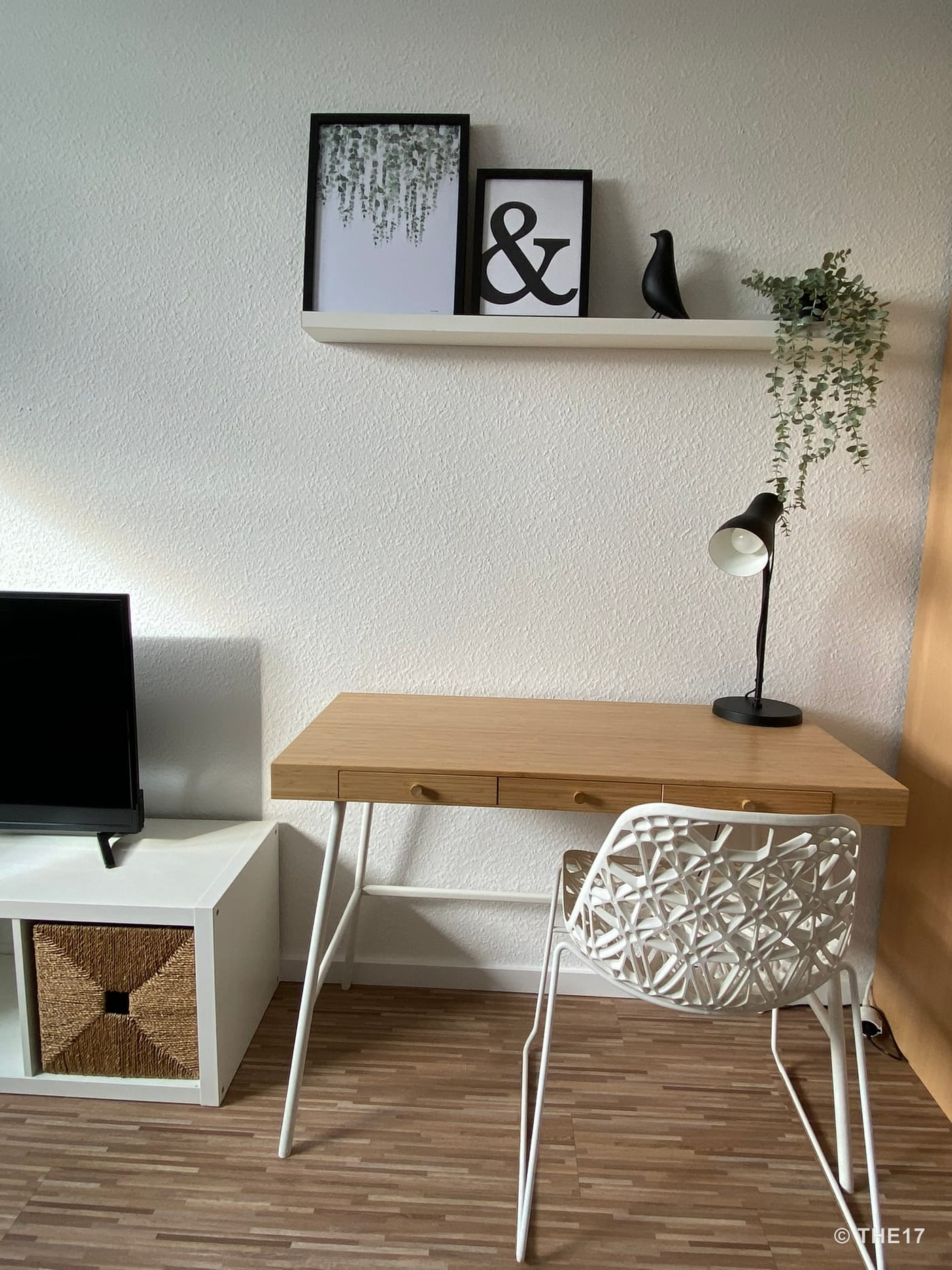 Schreibtisch mit drei Schubladen, weißer Dekostuhl, schwarze Lampe und Regalfach mit Bildern und Pflanze an weißer Wand.