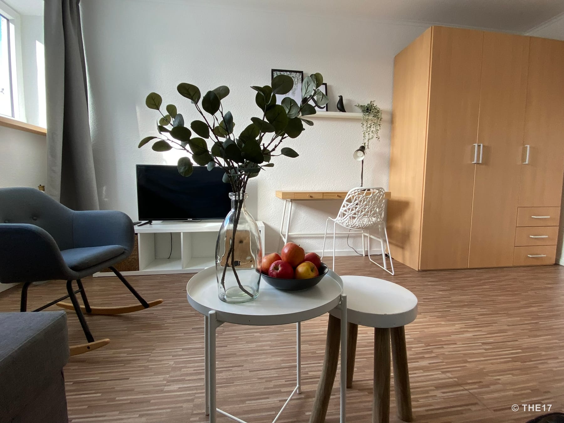 Wohnzimmer mit grauem Schaukelstuhl, kleinem weißen Couchtisch, TV, Schreibtisch mit Stuhl und großem Holzkleiderschrank.