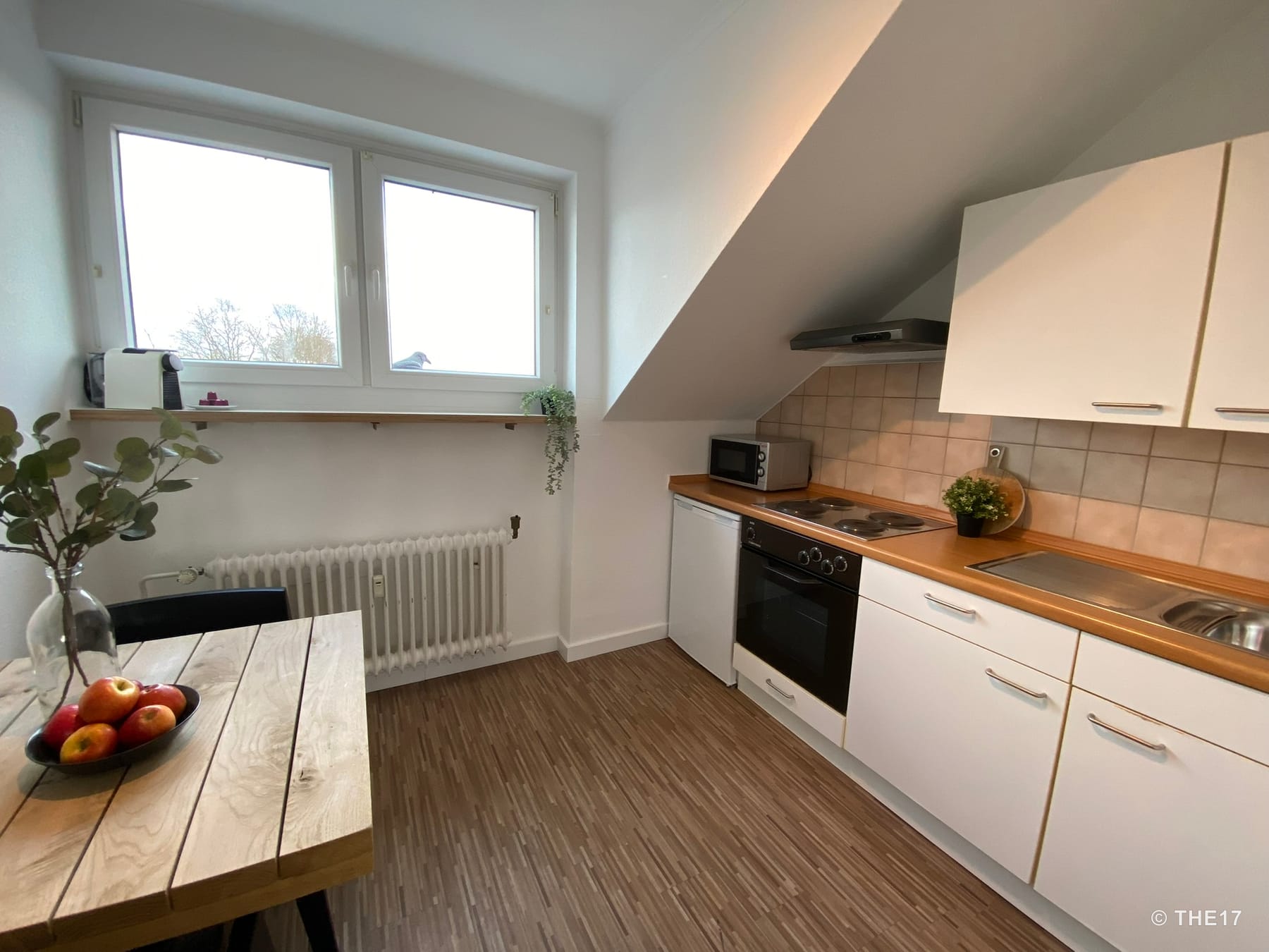 Kleine Küche mit weißer Schränken, Holzarbeitsplatten, einem Herd, Mikrowelle, Hohem Fenster und Holz-Esstisch mit Obstschale.