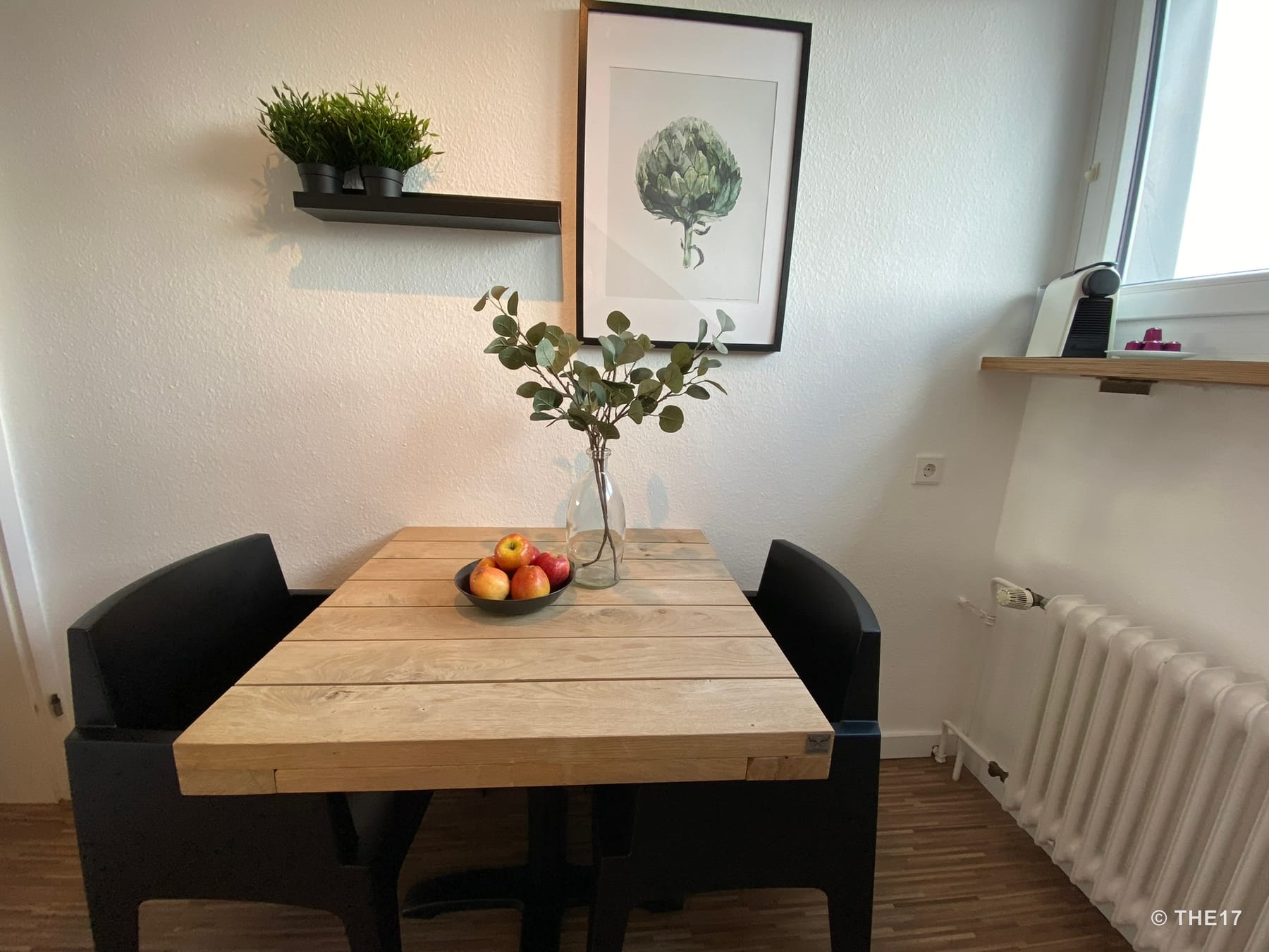 Kleiner Esstisch aus Holz mit zwei schwarzen Stühlen, Obstschale und Vase mit Zweigen, weiße Wand mit Bild und Pflanzenregal.