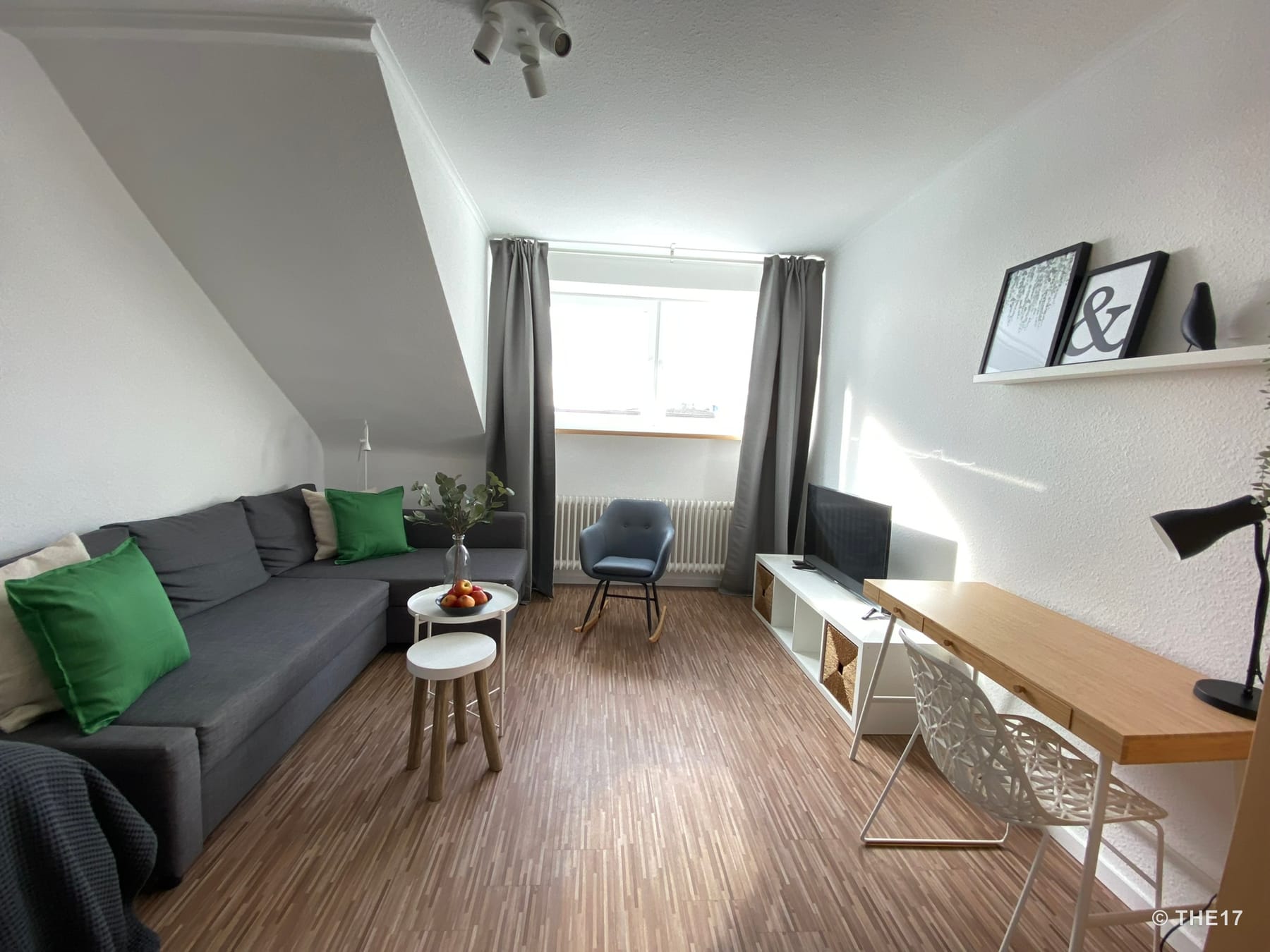 Wohnzimmer mit grauem Ecksofa, grün-beigen Kissen, kleinem runden Tisch, blauem Schaukelstuhl, Schreibtisch mit Weißem Stuhl, TV und Dekoration.