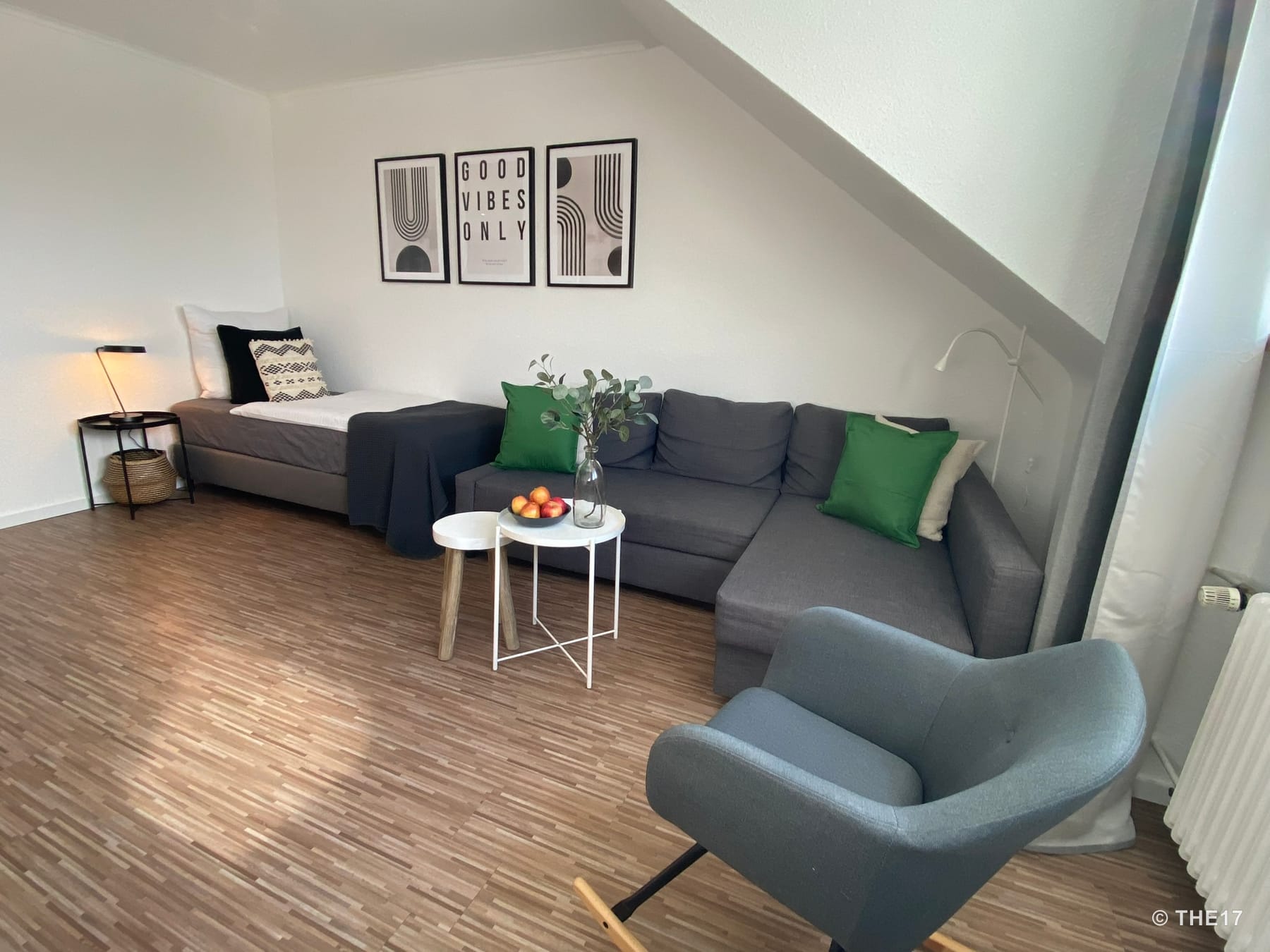 Wohnzimmer mit grauem Sofa, grüne Kissen, grauer Sessel, Einzelbett mit Nachttisch und moderner Wandkunst.