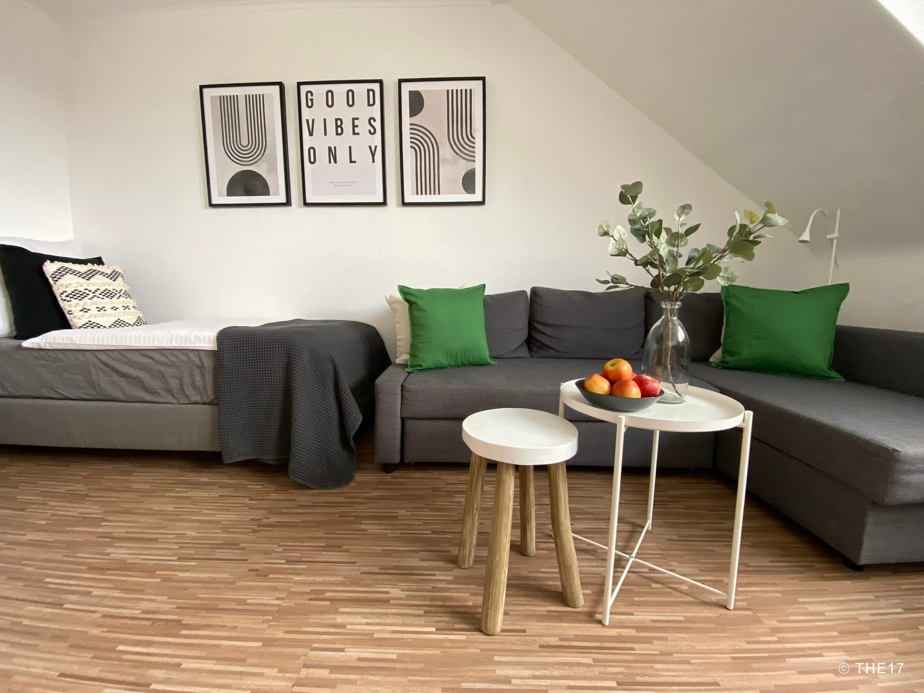 Wohnzimmer mit grauem Ecksofa, grünem Kissen, Einzelbett, drei Wandbildern und kleinem weißen Tisch mit Obstschale und Vase.