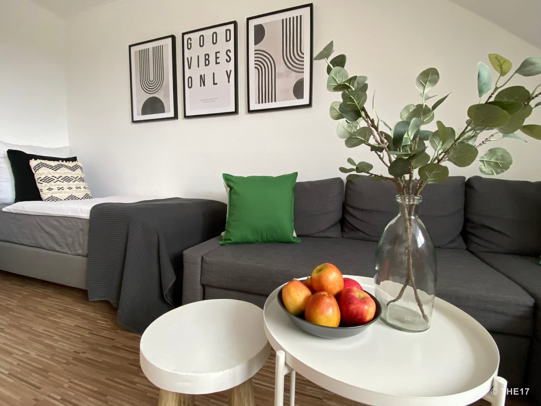 Living room with gray sofa, green pillow, side table with apples and vase, white walls with three framed pictures.