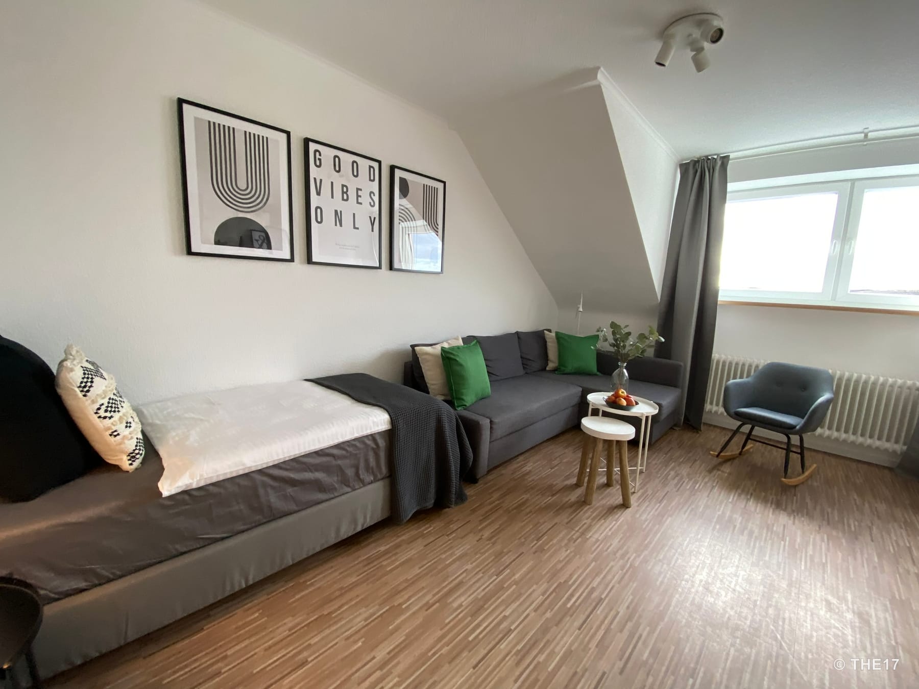 Wohnzimmer mit grauem Ecksofa, Tagesbett, kleinem Beistelltisch, Schaukelstuhl und drei modernen Bildern an der Wand.