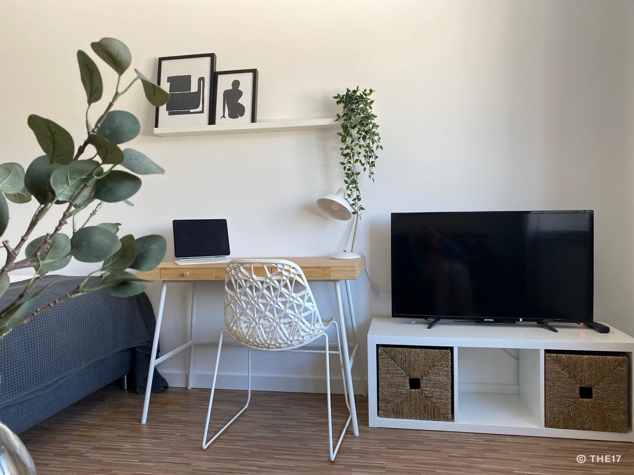 Schreibtisch mit Laptop, weißem Stuhl und Pflanze. Fernseher auf weißem Lowboard mit zwei Korbschubladen, Bett mit dunkelgrauer Decke.
