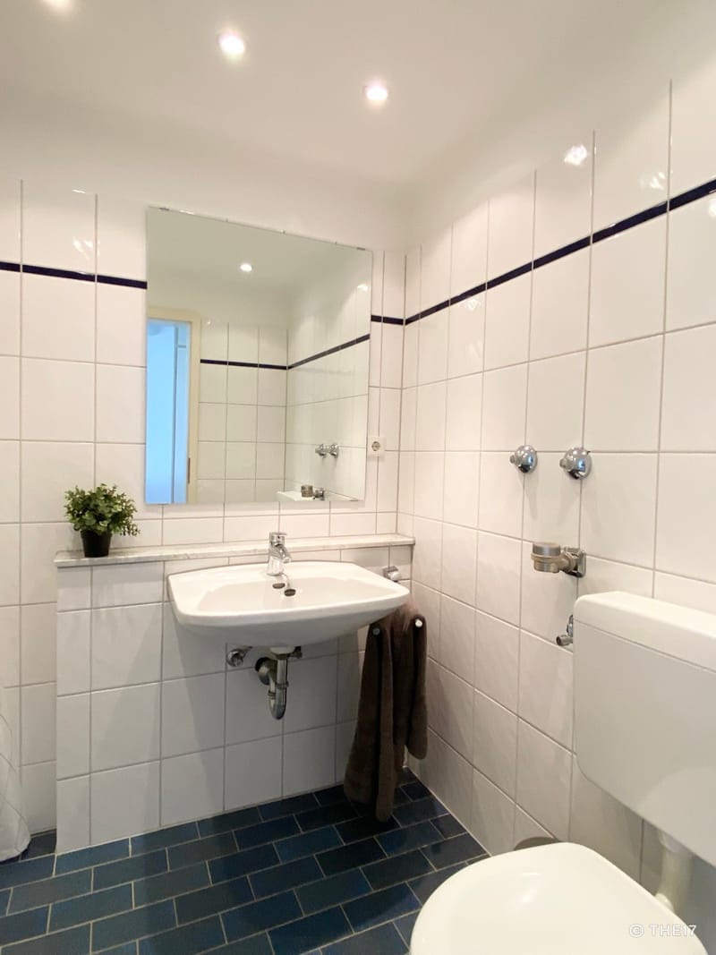 Helles Badezimmer mit weiß gefliesten Wänden, blauem Boden, weißem Waschbecken, Spiegel und grauem Handtuch.
