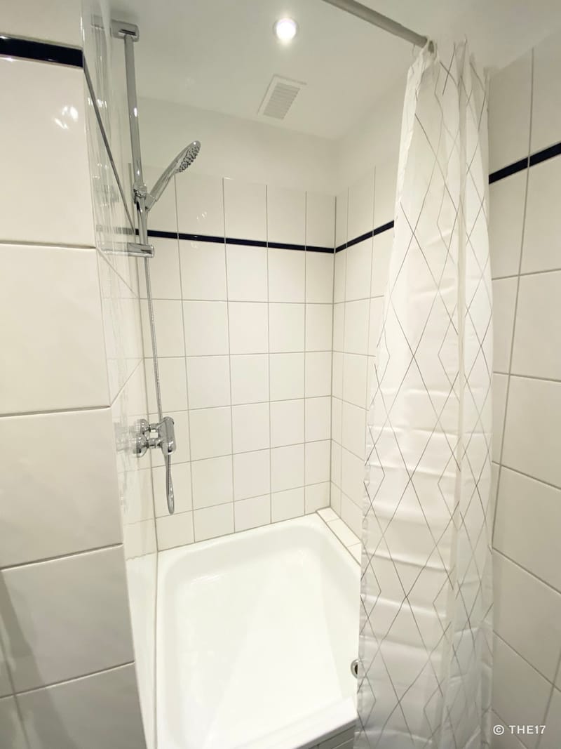 Weiße Duschkabine mit Chrom-Duschkopf, weißen Kacheln und weißem Duschvorhang mit grauem Muster.