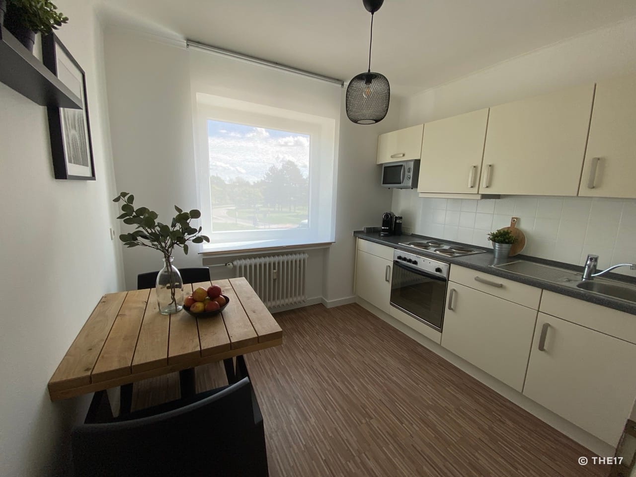 Kleine helle Küche mit weißem Schrank, Holz-Tisch, zwei schwarzen Stühlen und großem Fenster mit Sicht auf Bäume.