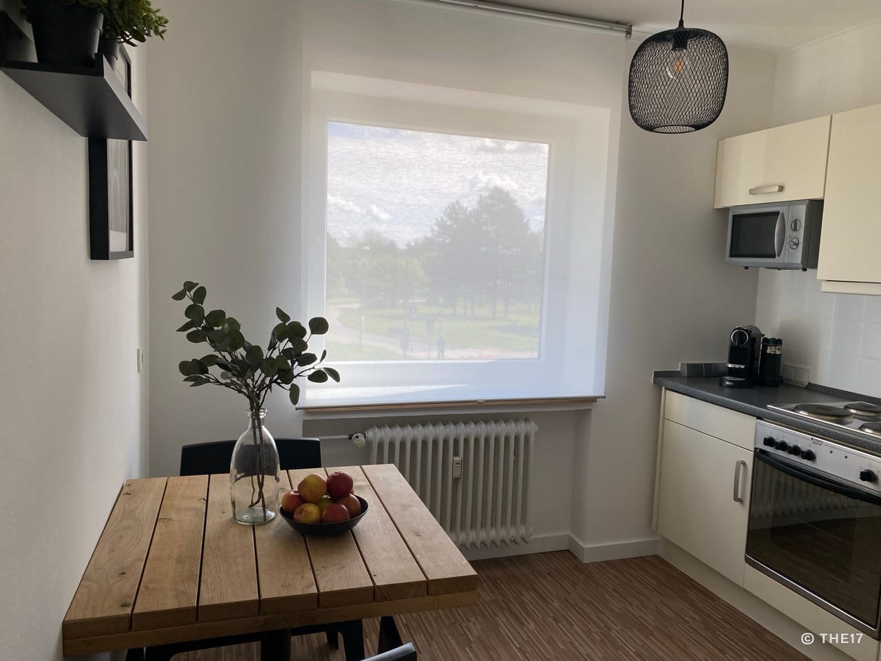 Kleine Küche mit Holztisch, Obstschale, Pflanzenzweig in Glasvase und großen Fenster mit weißem Vorhang.