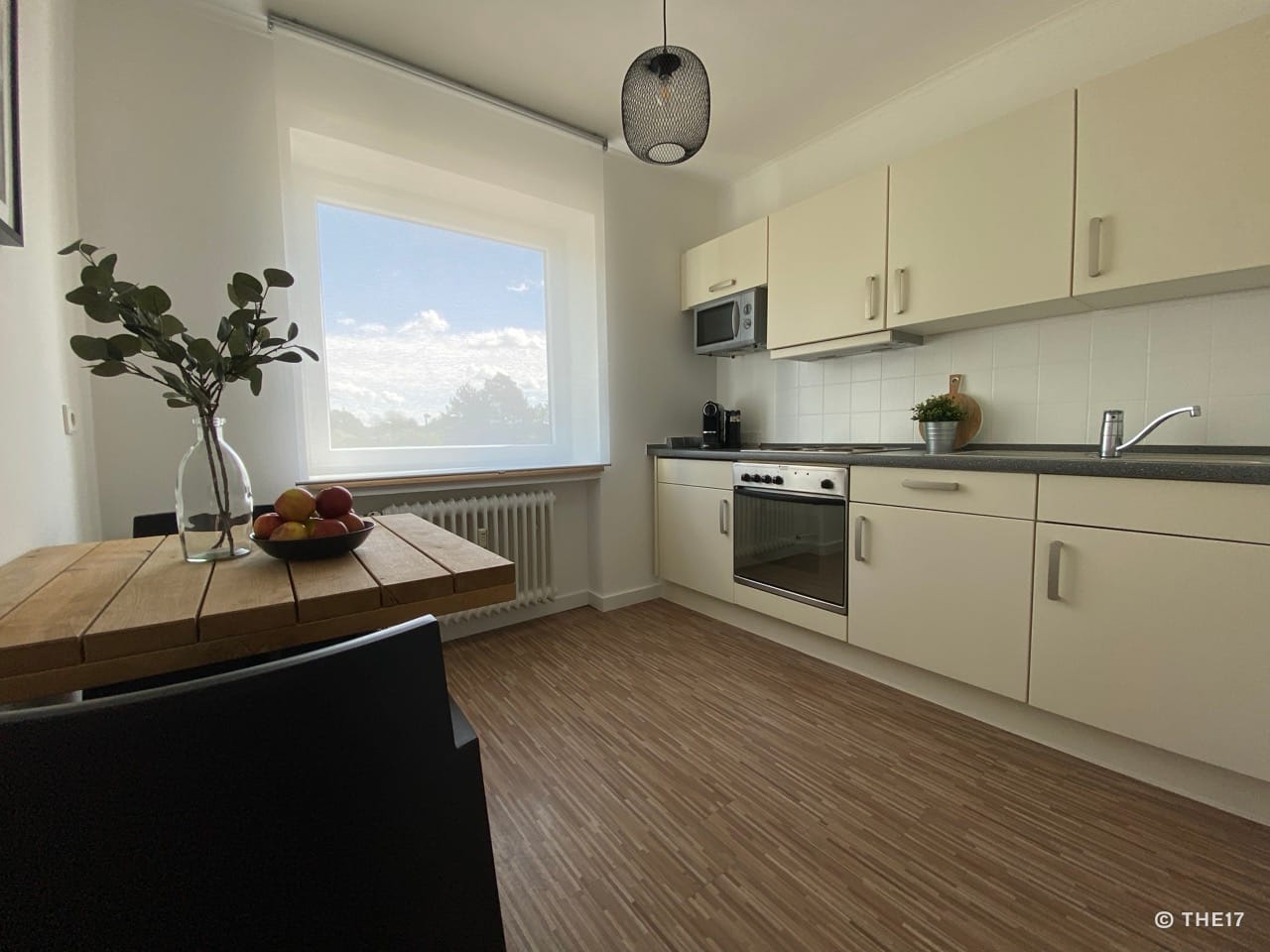 Kleine Küchenzeile mit hellen Schränken, Backofen, Mikrowelle und Holztisch neben einem Fenster mit Blick nach draußen.