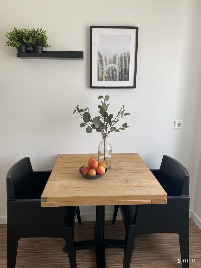 Holztisch mit zwei schwarzen Stühlen, Schale mit Äpfeln und Vase mit Zweig, Wandbild mit Kaktus und kleines Regal mit Pflanzen.