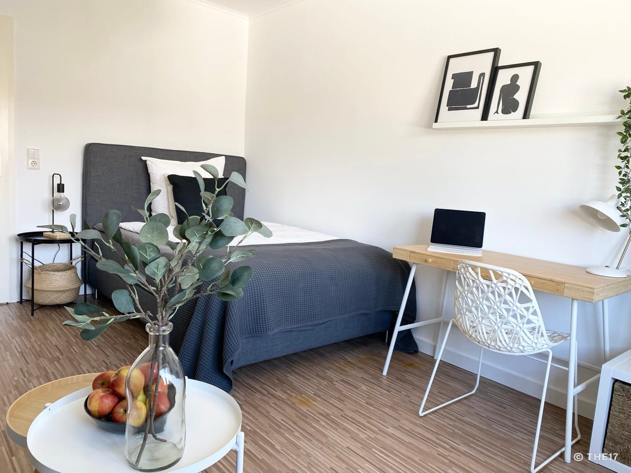 Schlafzimmer mit grauem Bett, Holzboden, Schreibtisch mit Laptop, weißem Stuhl und Pflanzen in der Vase im Vordergrund.