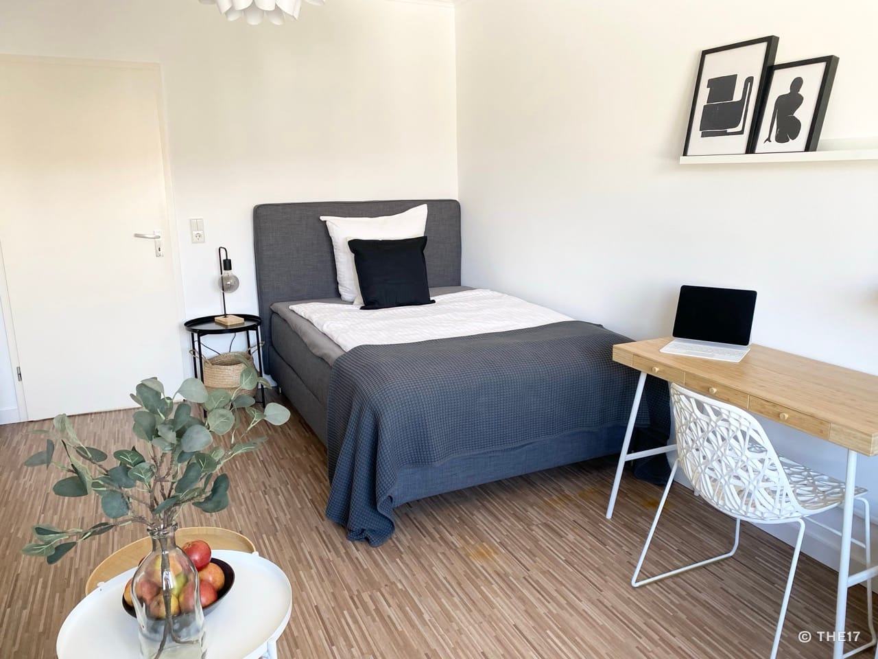 Einzelzimmer mit grauem Bett, Holzschreibtisch, weißem Stuhl, Nachttisch mit Lampe und einfacher Wanddekoration.