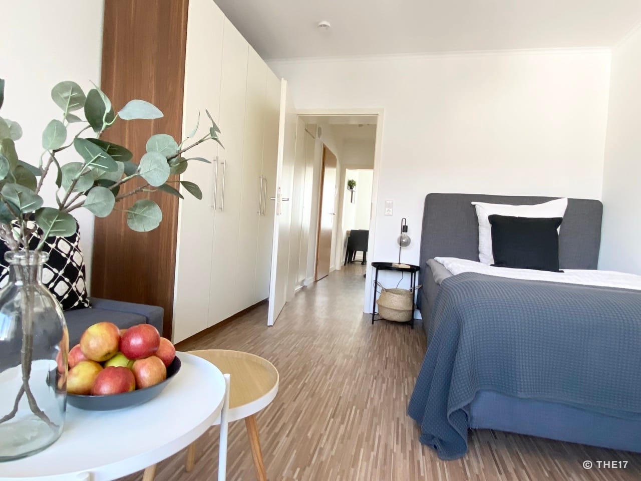 Schlafzimmer mit grauem Bett, weißen und schwarzen Kissen, weißen Wandschrankfronten und Holzboden.