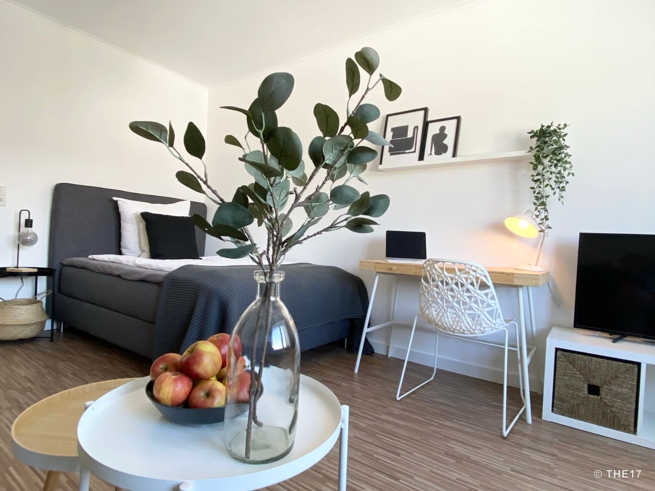 Modern eingerichtetes Zimmer mit grauem Bett, Schreibtisch, Stuhl, TV und dekorativem Tisch mit Vase und Äpfeln.